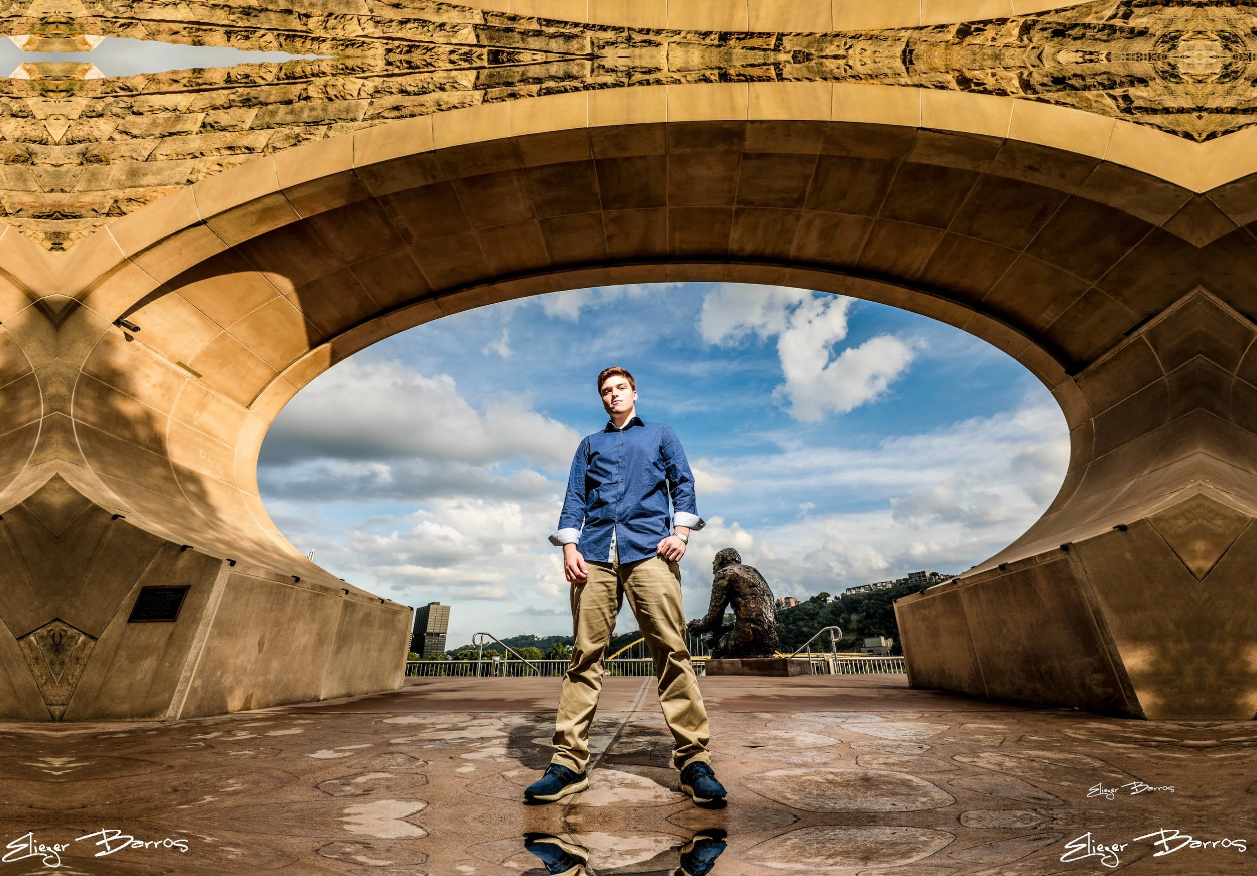 A young man standing beneath a large, modern, circular arch structure made of stone and concrete, against a backdrop of blue sky with clouds and distant cityscape.