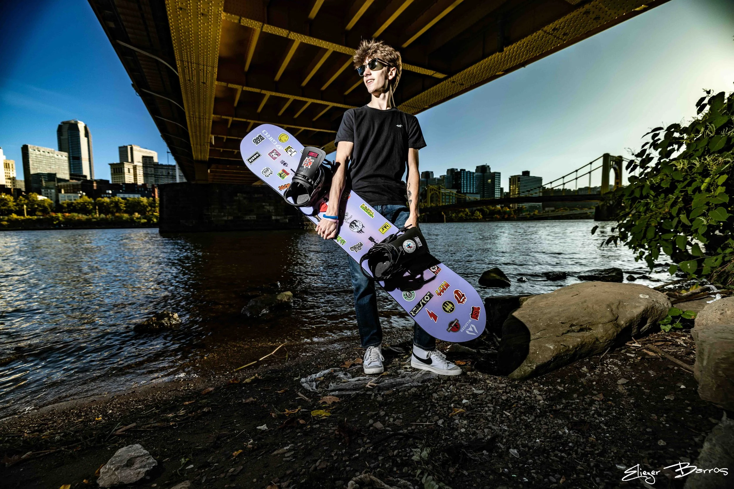 Young man wearing sunglasses and black t-shirt holding a snowboard with stickers, standing on rocks by a river with cityscape and bridge in the background.
