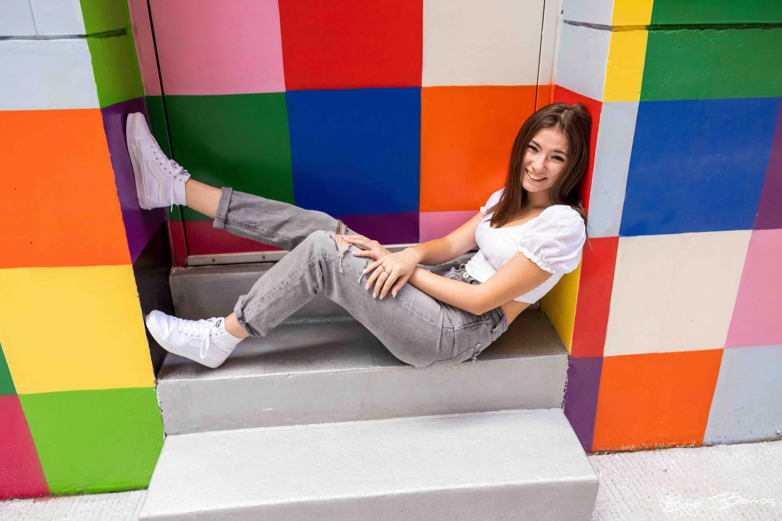 A young woman is sitting on colorful painted stairs with a smile, wearing white sneakers, gray ripped jeans, and a white top, in front of a multicolored wall with blocks of red, green, yellow, purple, orange, and pink.