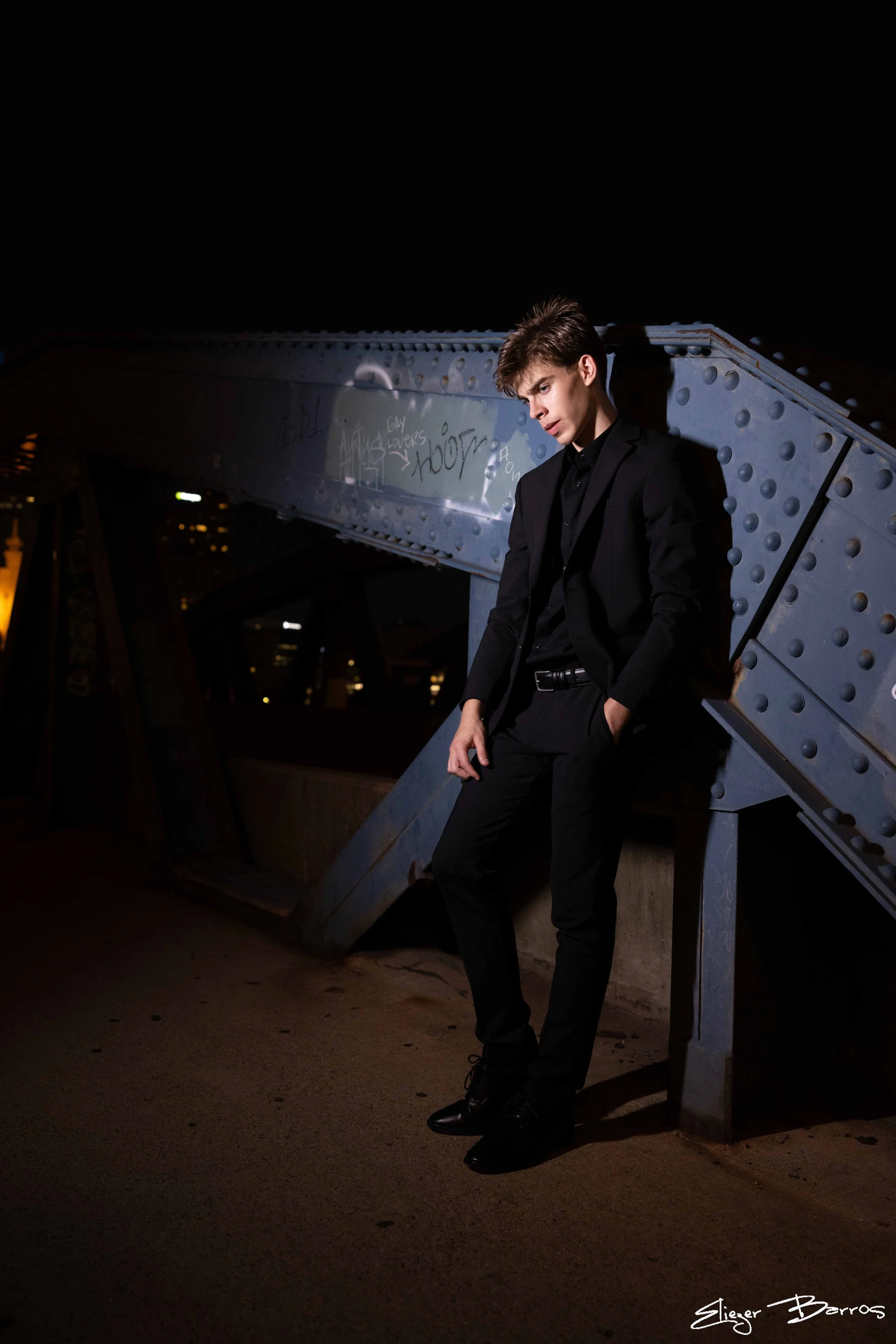 A young man in a black suit and shoes leaning against a metal bridge at night, with graffiti on the bridge and city lights in the background.