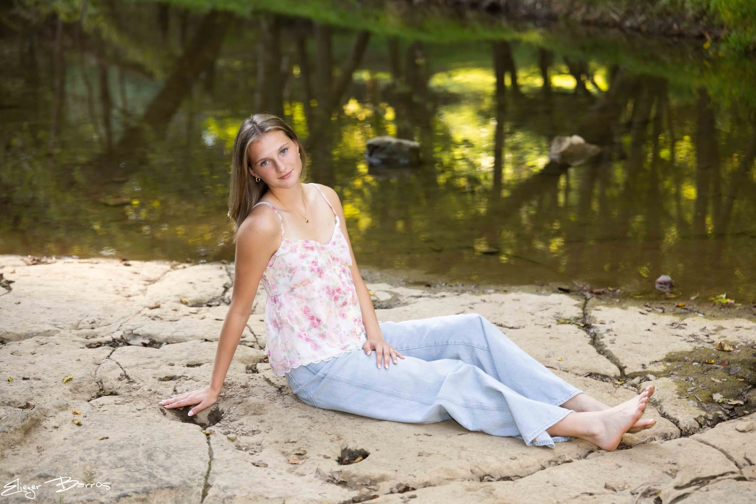A young woman with long light brown hair wearing a pink floral sleeveless top and light blue jeans sitting on rocks by a wooded riverbank, looking at the camera with a gentle expression.