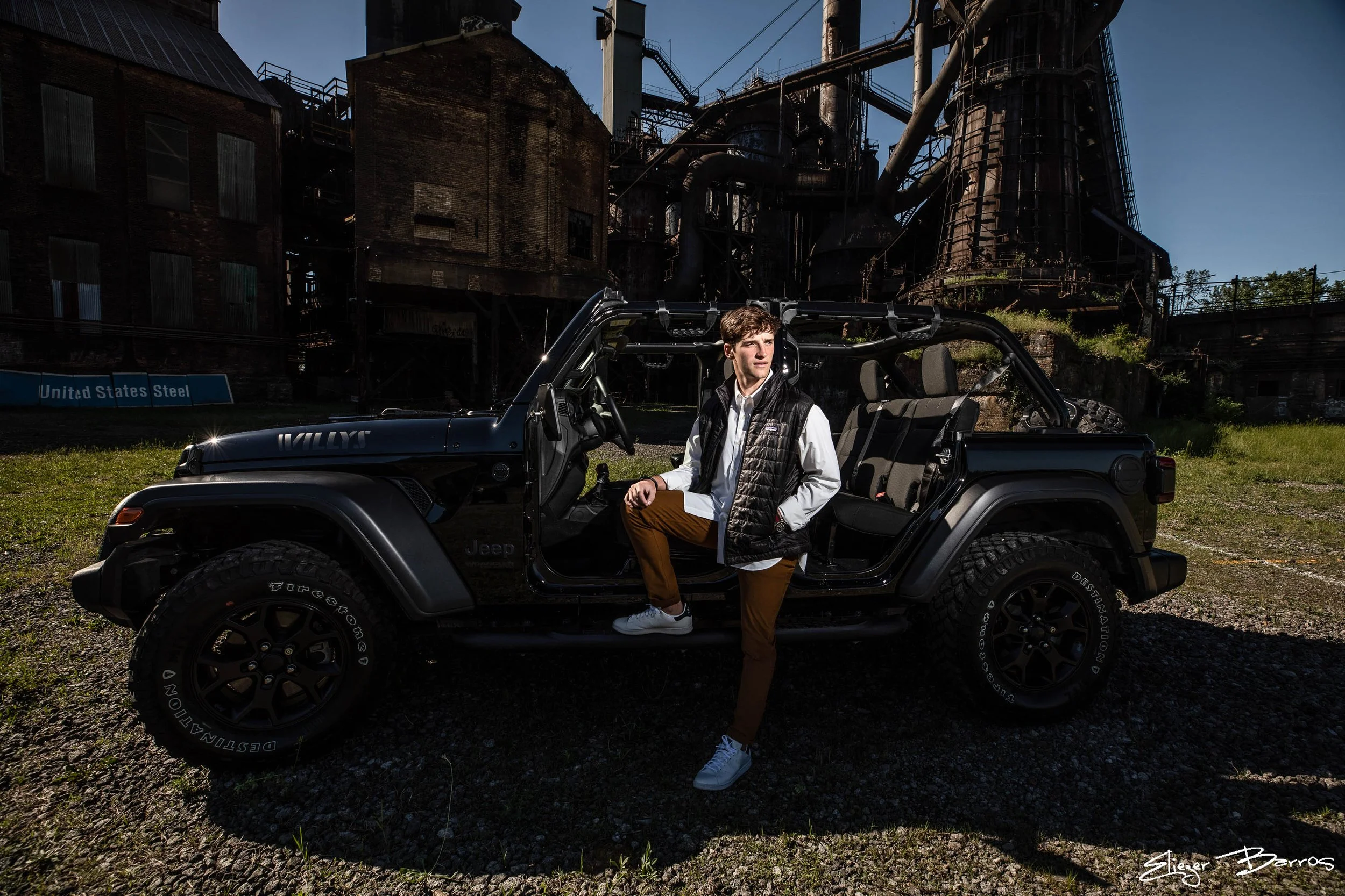 A young man with brown hair stands next to a black Jeep Wrangler, partially seated on the door frame, in front of an old industrial factory with rusted structures and pipes. The man is wearing a white shirt, black vest, and brown pants, with white sn