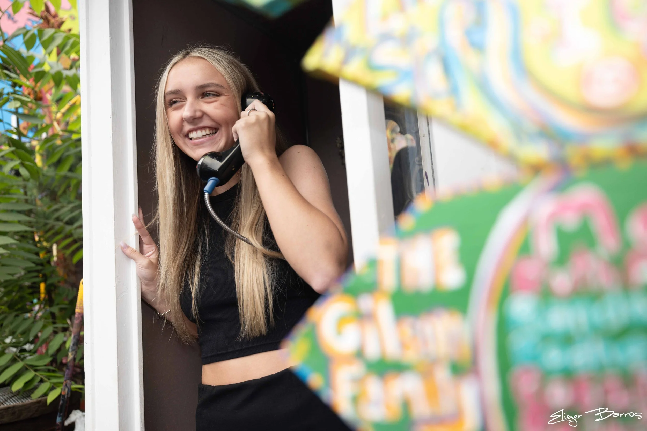 A young woman with long blonde hair is smiling while talking on a black phone, standing in a small wooden booth with greenery visible outside.