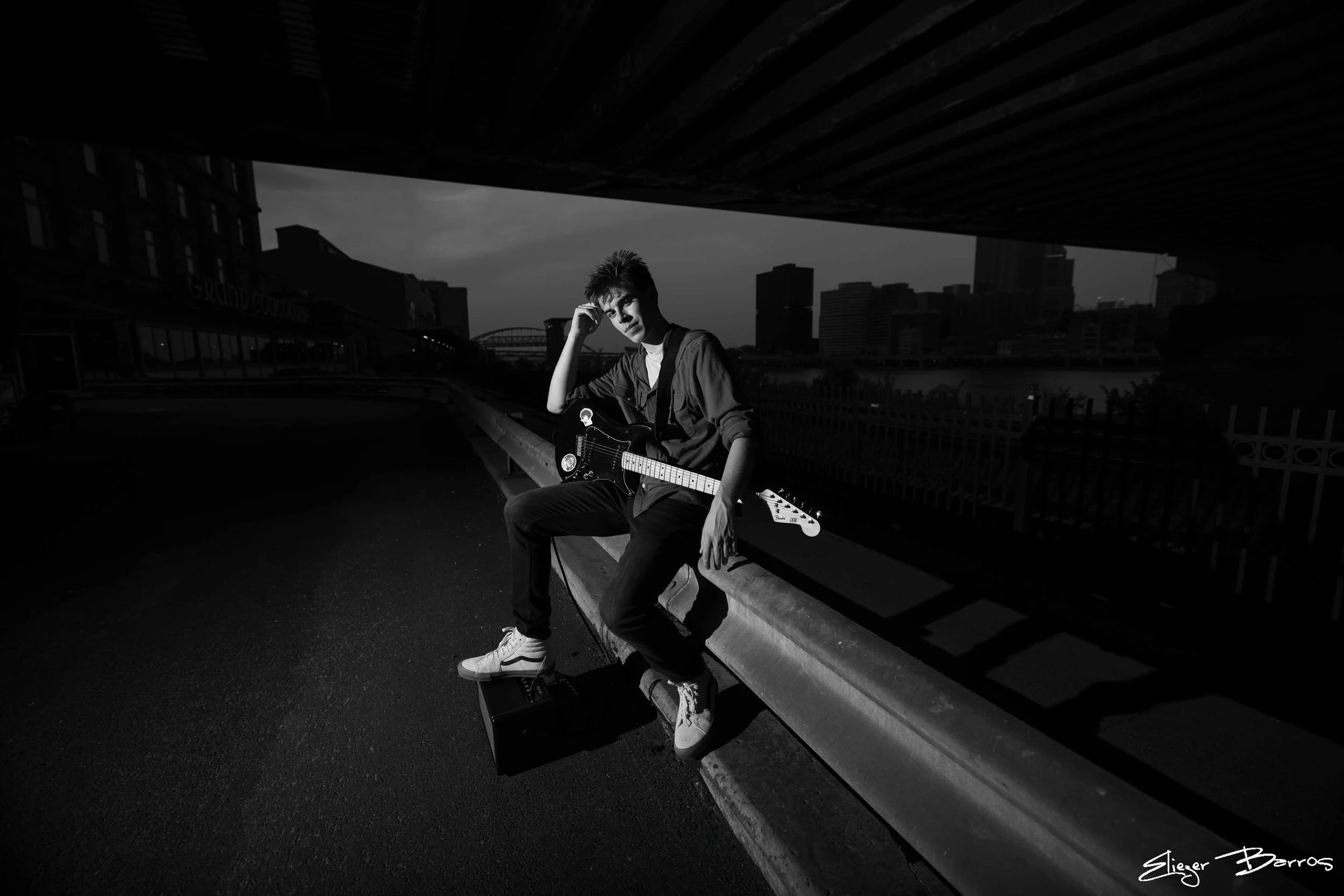 A young man sitting on a park bench under an overpass, holding an electric guitar, with a city skyline in the background, during daytime.