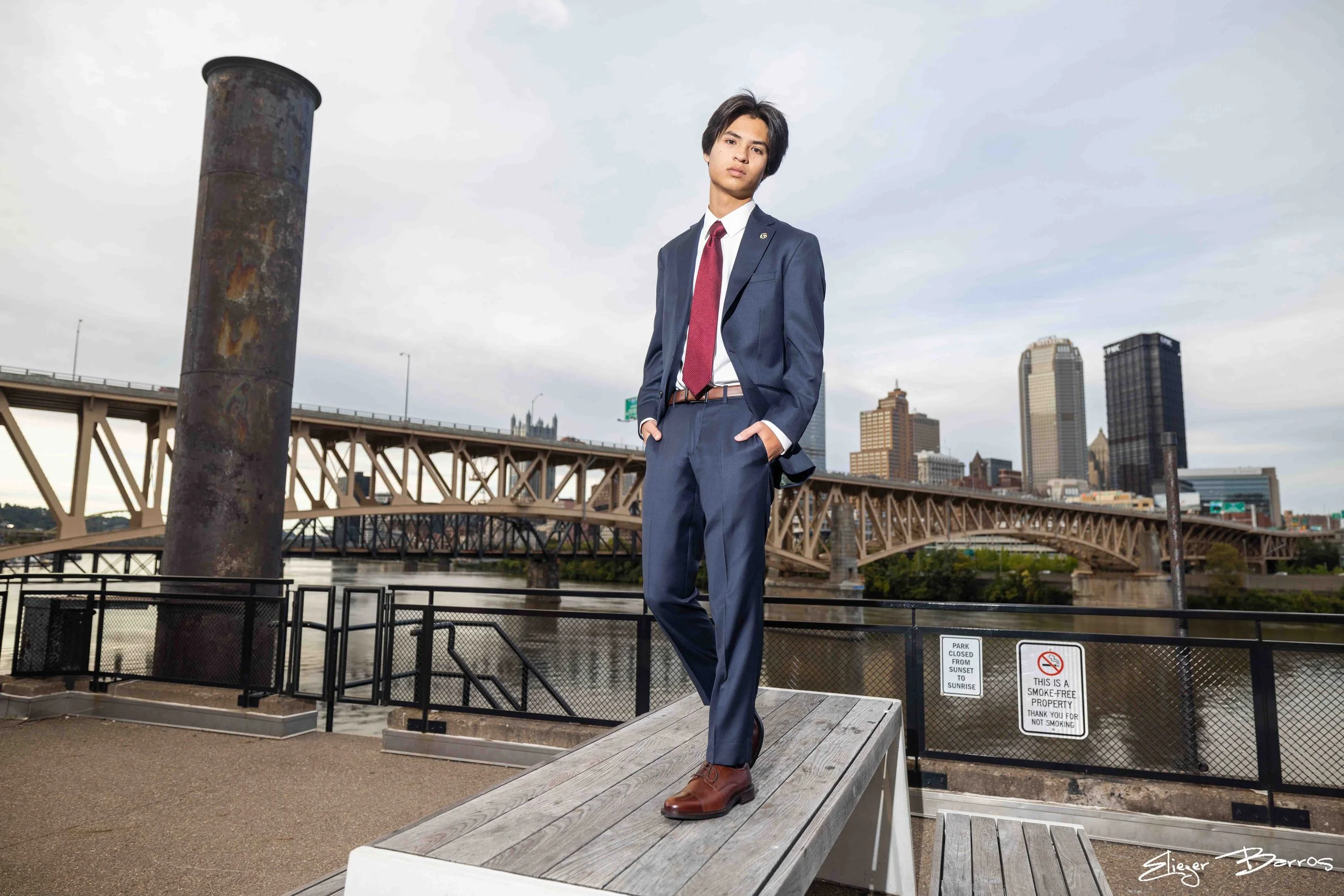 Senior Pictures Pittsburgh at Highline & Color Park. Young man in a blue suit posing on a wooden overlook with river, bridges, and city skyline backdrop during his stylish urban senior photoshoot.