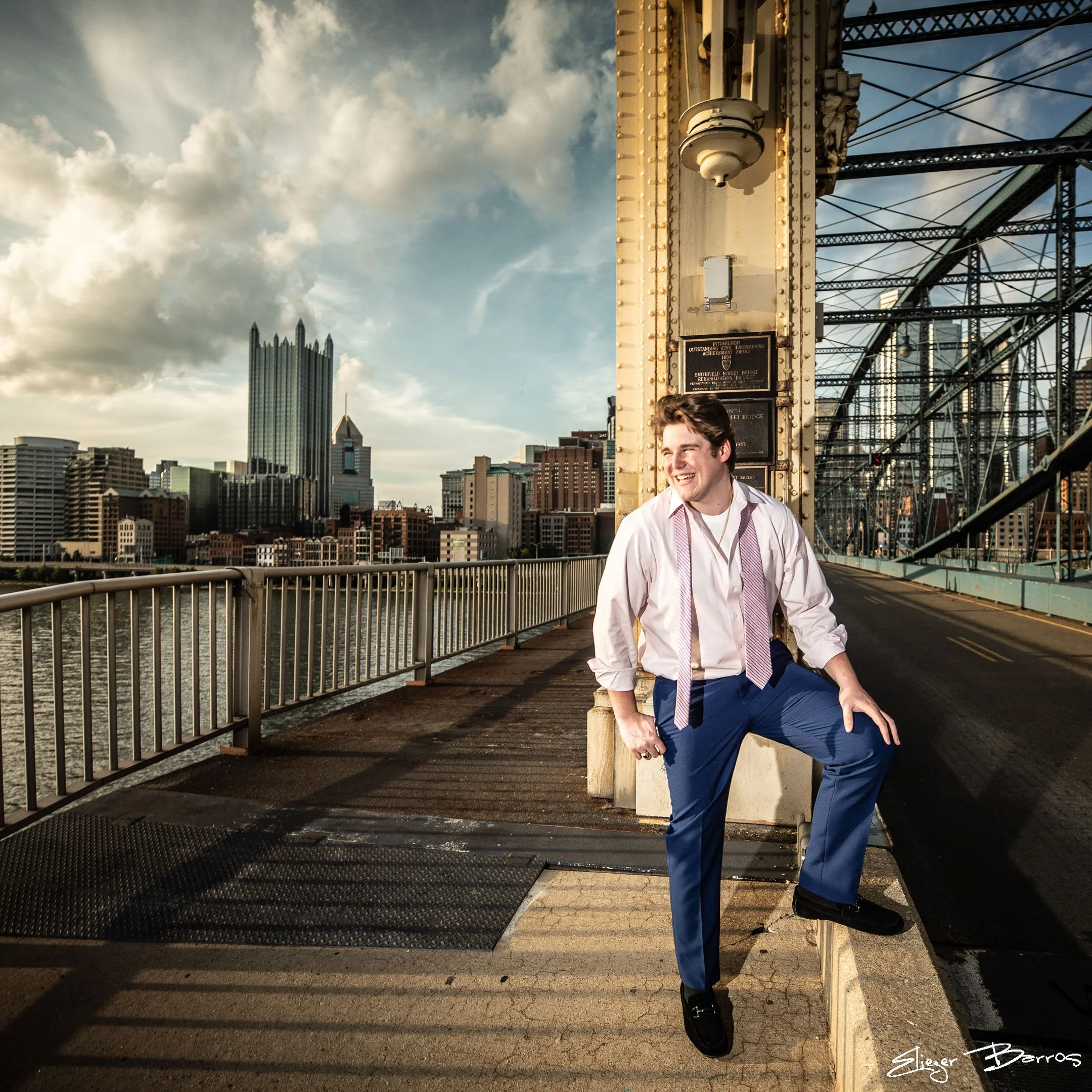 A young man in a white shirt with purple tie and blue pants, smiling and leaning against a concrete post on a bridge with city buildings in the background, during sunset or late afternoon.