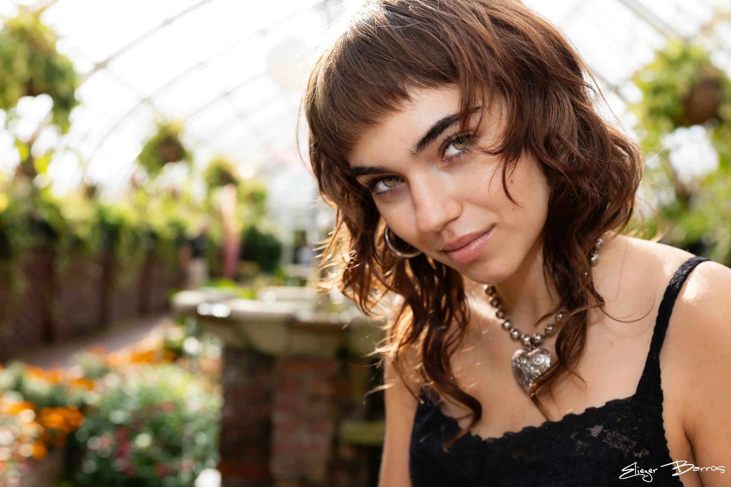 A woman with curly brown hair and green eyes looking at the camera outdoors with greenery and flowers in the background.