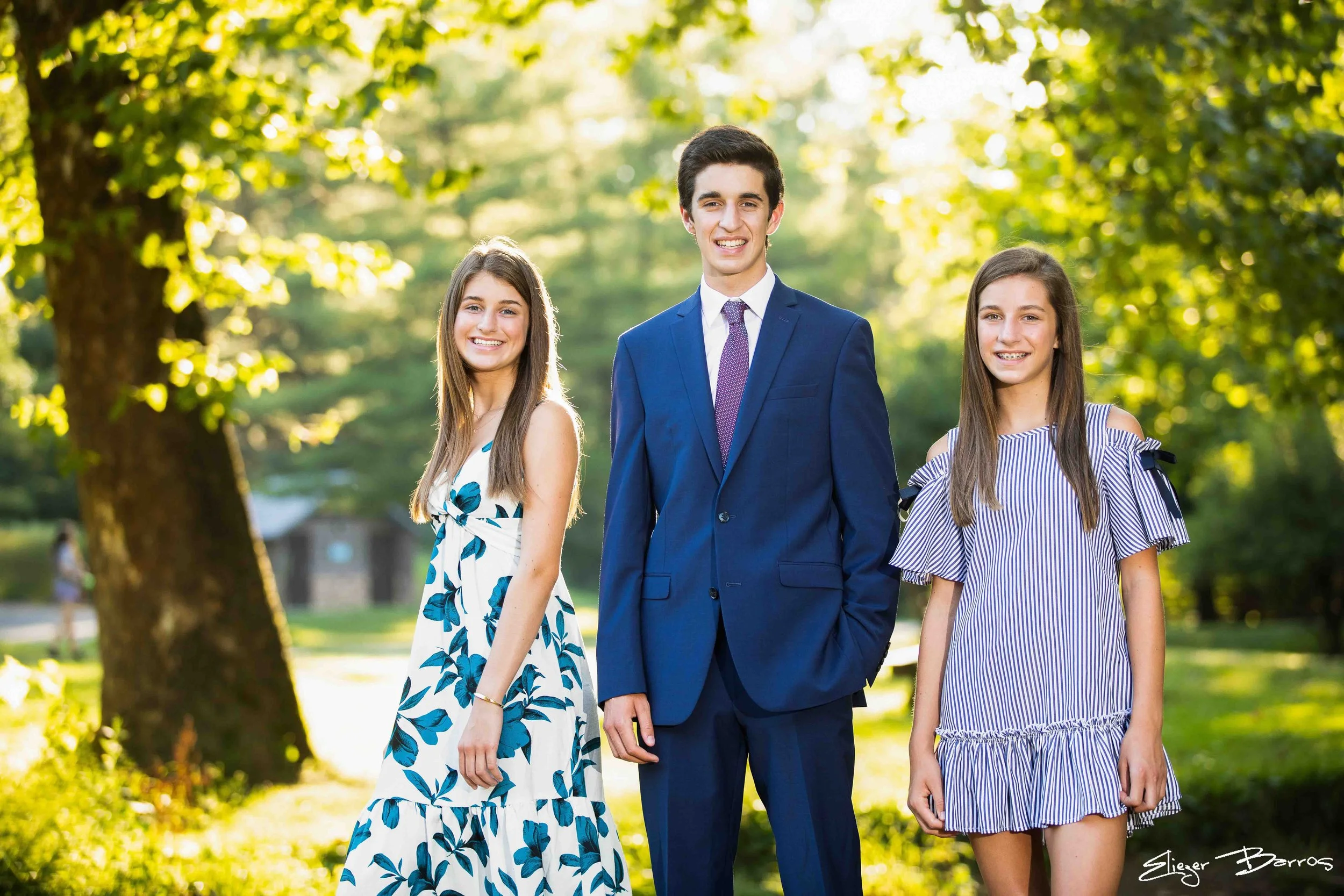 Three teenagers, two girls and one boy, walking outdoors in a park with green trees in the background, smiling at the camera.