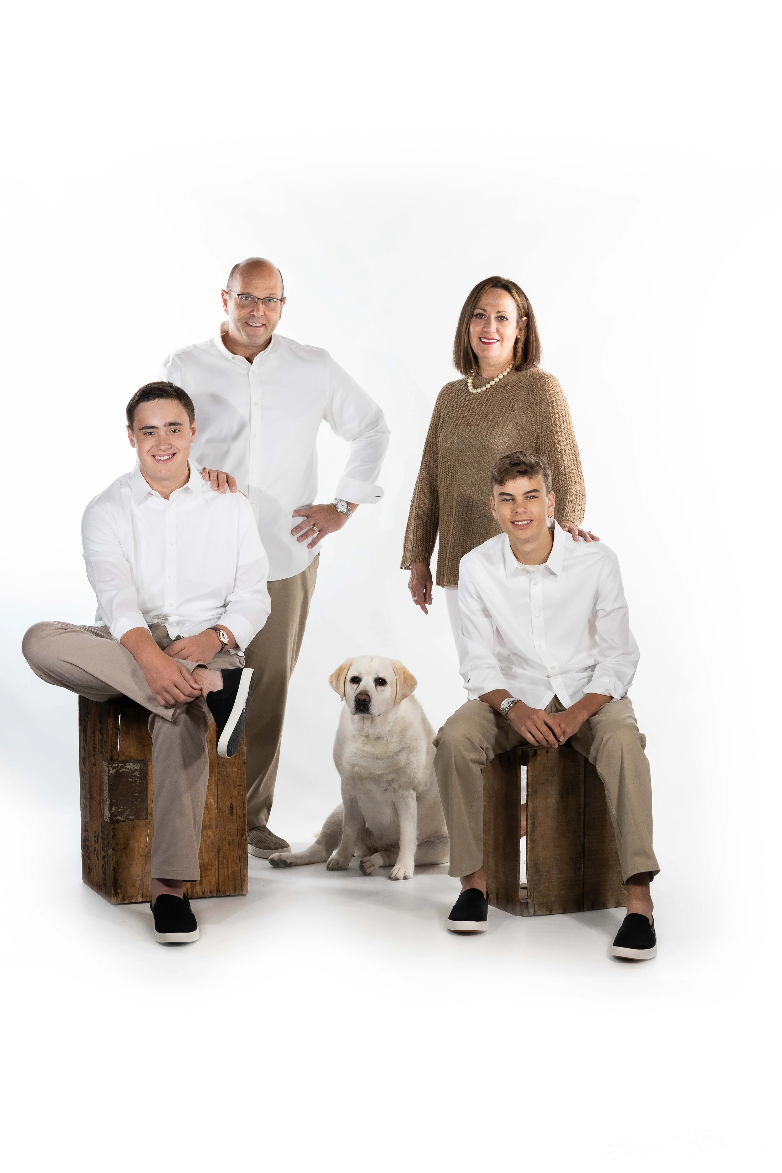 A family of five people and a dog posing against a white background. The family includes an older man and woman, and three young males. The older man is standing with one hand on his hip, the woman is standing and smiling, and the two young males are