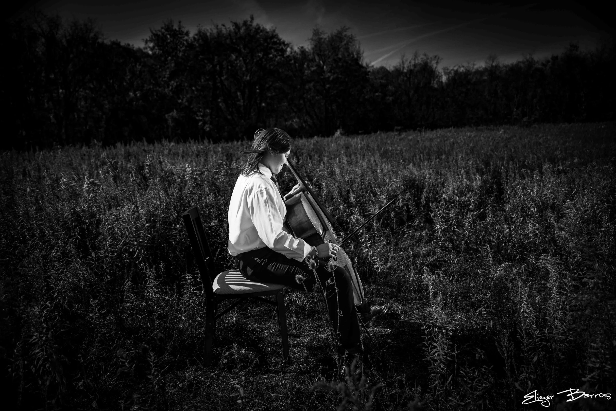 A young girl sitting outdoors in a field playing a cello at dusk, with trees and a darkening sky in the background.