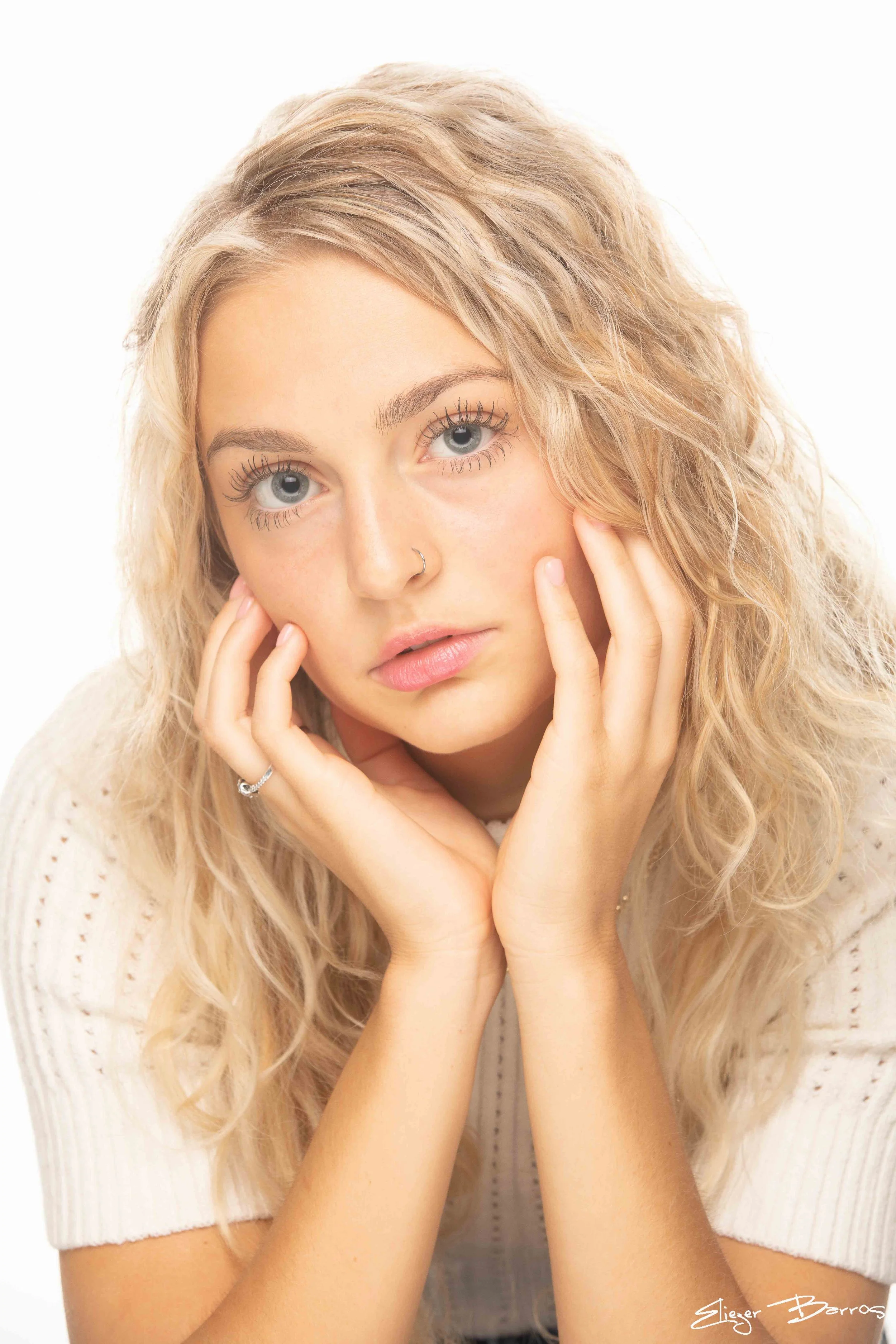 A young woman with long curly blonde hair, blue eyes, and light skin, touching her face with her hands, wearing a white knitted top and a ring on her finger, against a white background.
