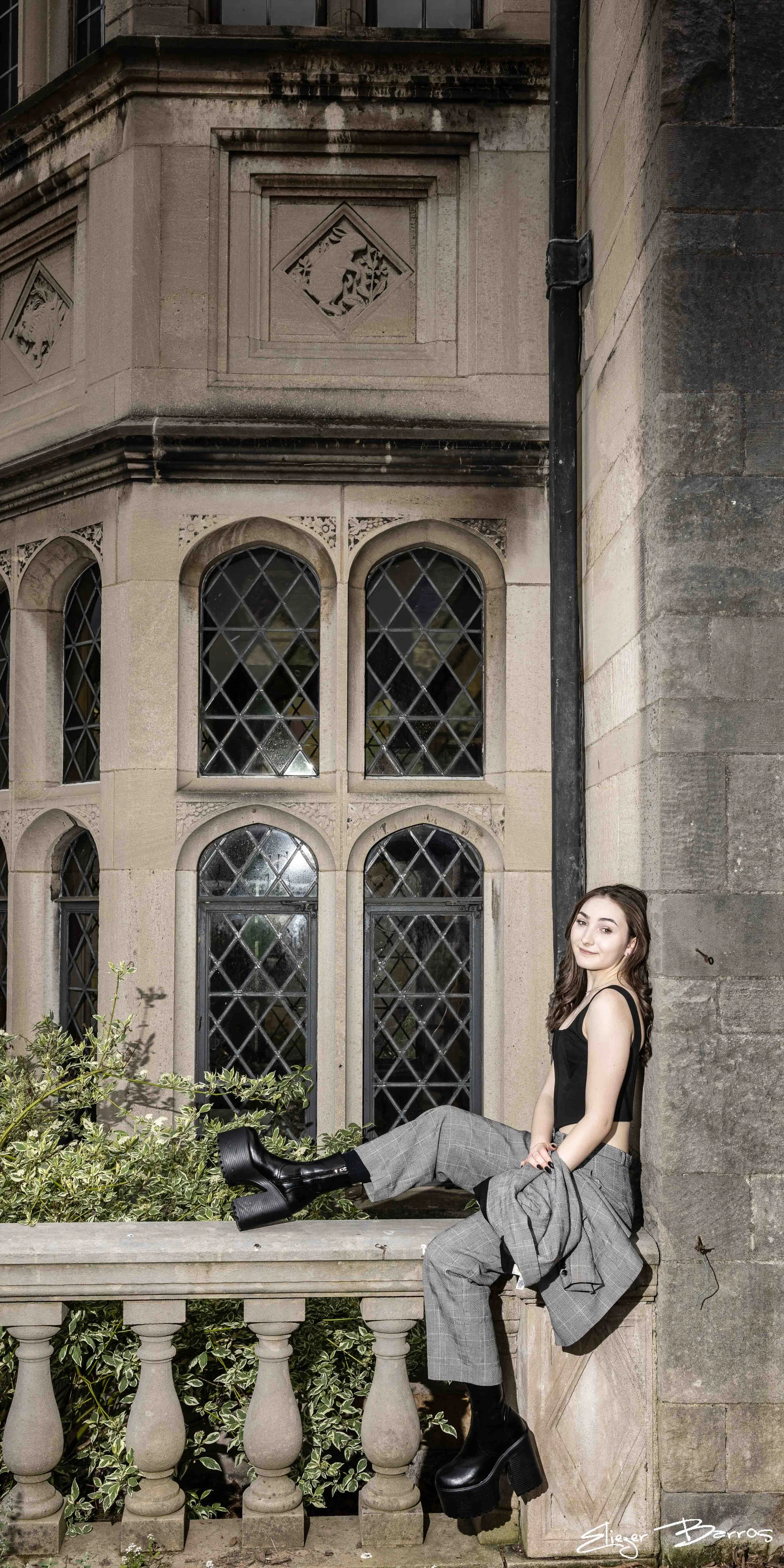 A young woman in black crop top and plaid pants sitting on a stone ledge outside a building with large, gothic-style windows and ornate stone detailing.