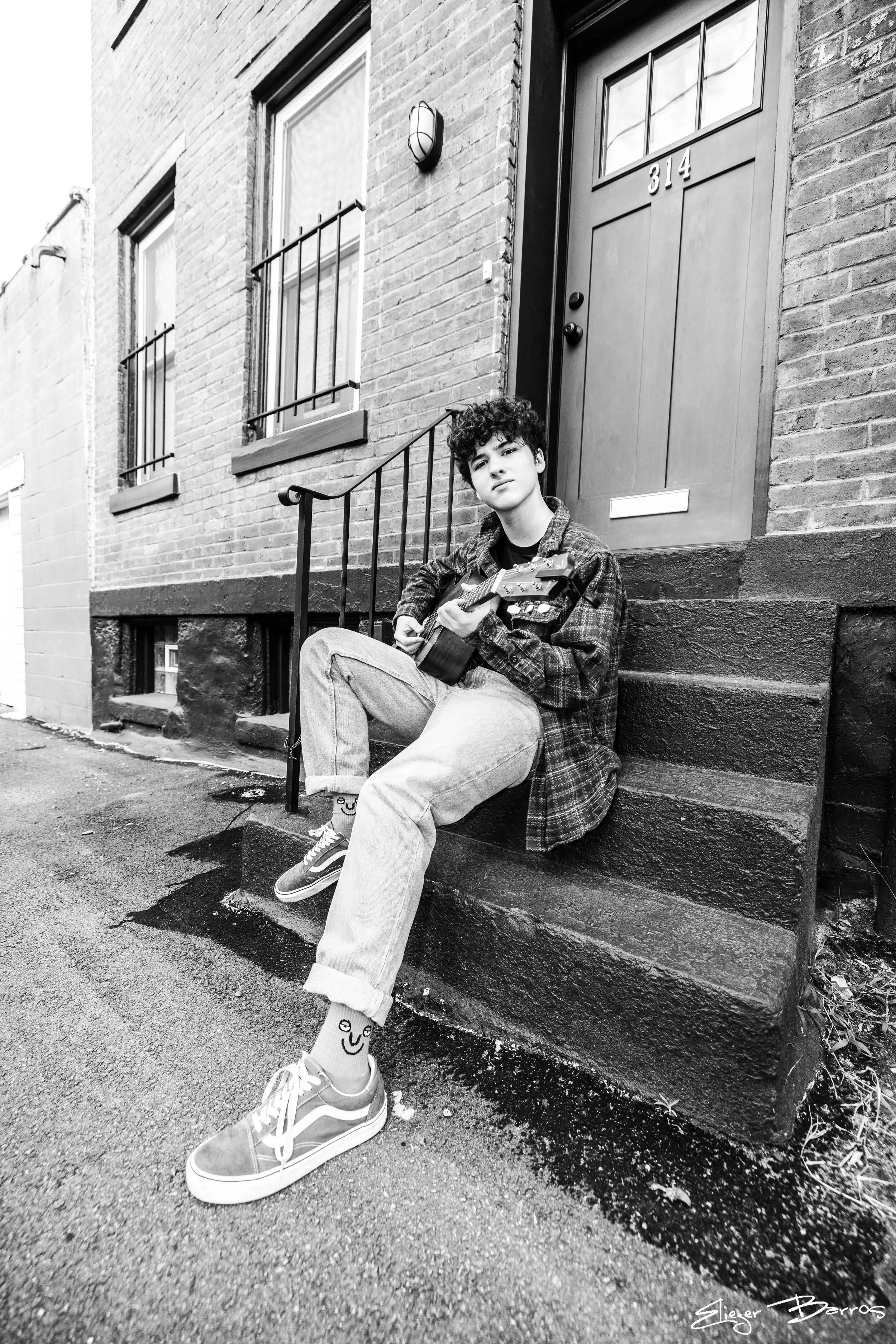 Black-and-white photo of a young man with curly hair sitting on the steps outside a brick building. He is holding a guitar, wearing casual clothes, sneakers, and patterned socks. The building has a front door with the number 314 and windows with bars
