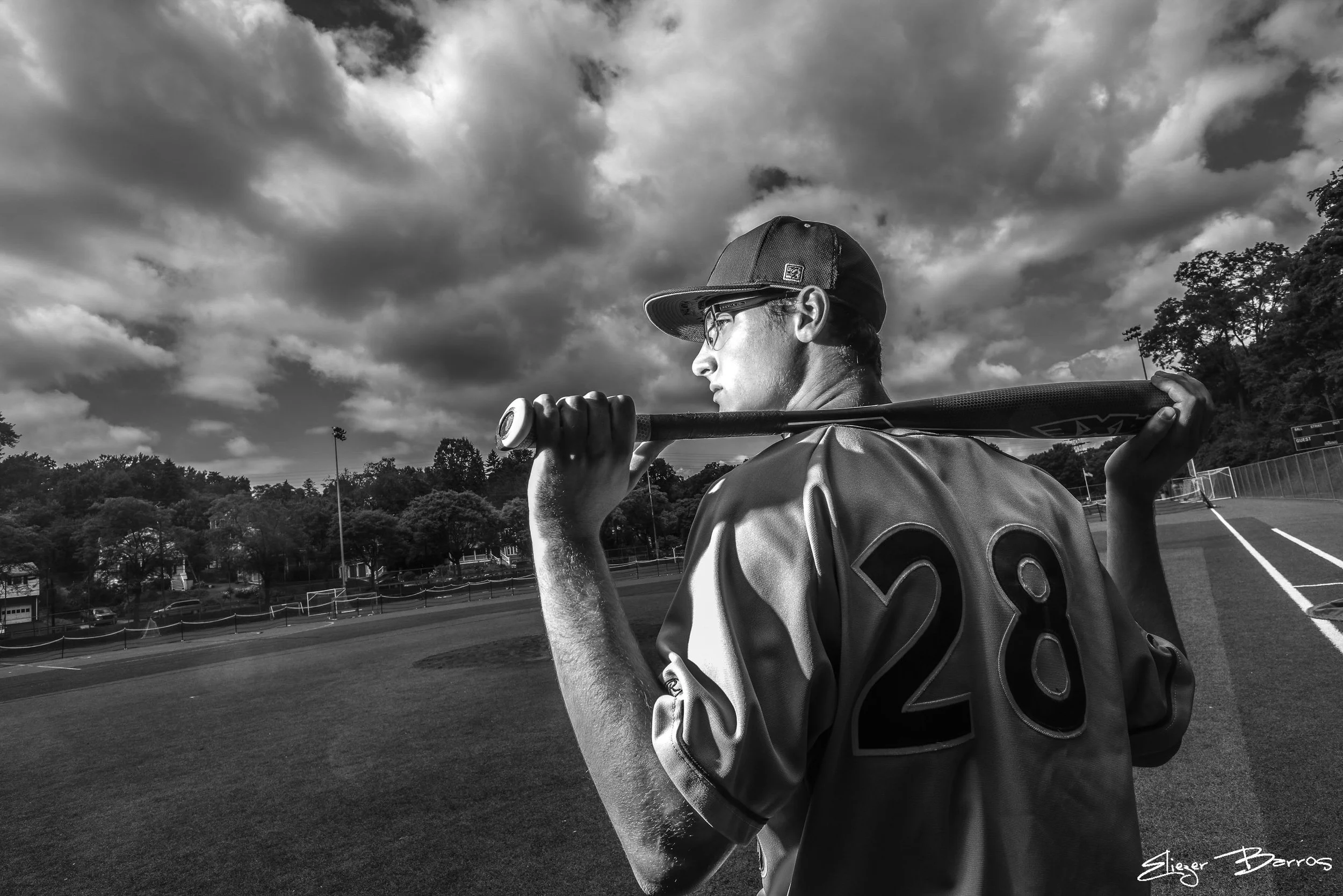 A black-and-white photo of a young man in a baseball cap and glasses, holding a baseball bat over his shoulder, standing on a sports field. The sky is cloudy and there are trees and a fence in the background.
