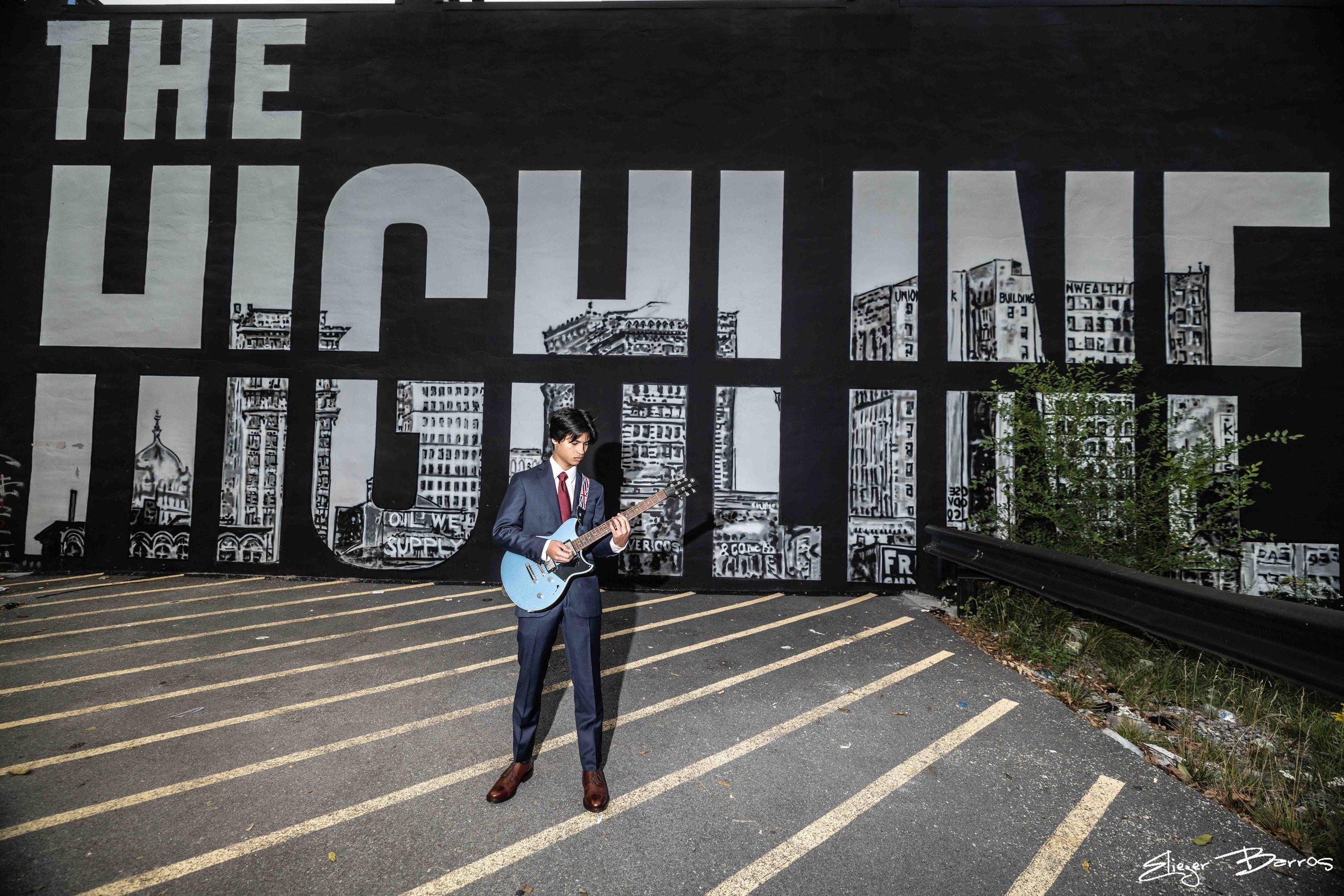 A young man in a suit playing an electric guitar outdoors on a street with painted cityscape wall behind him.