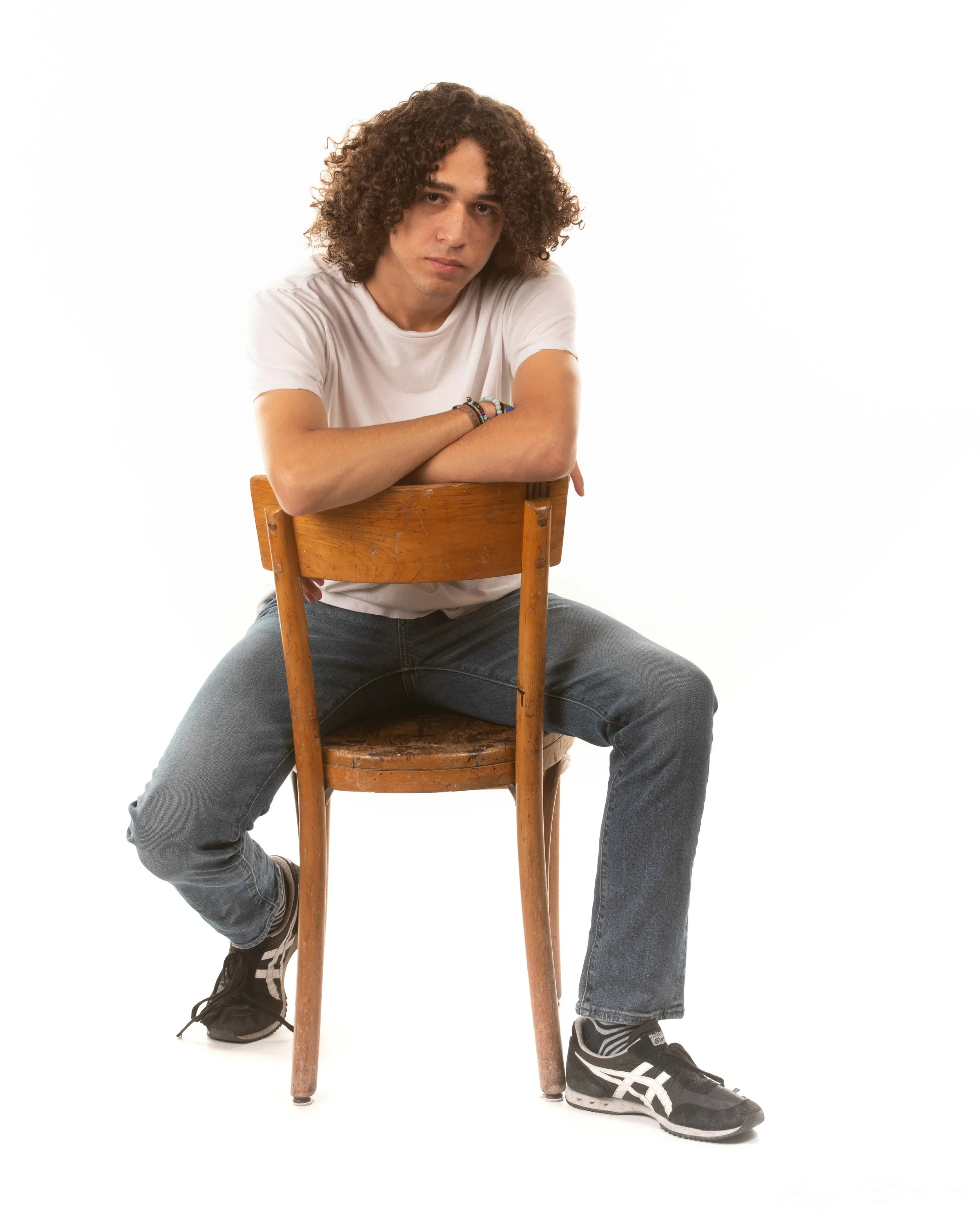 Young man with curly hair sitting on a wooden chair, leaning on the backrest with arms crossed, wearing a white t-shirt, gray jeans, and black sneakers, with a neutral expression