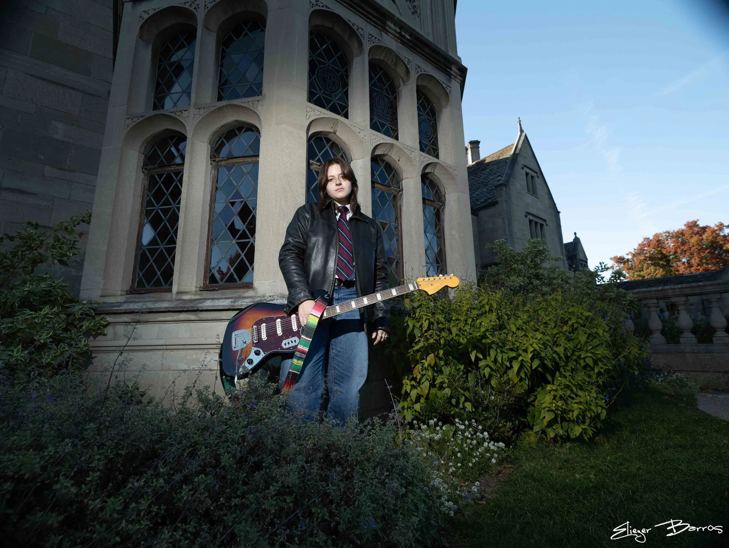 A young woman with dark hair, wearing a leather jacket, striped shirt, and jeans, stands outdoors in front of an old stone building with large arched windows. She holds an electric guitar with a rainbow strap. There are plants and shrubbery around he