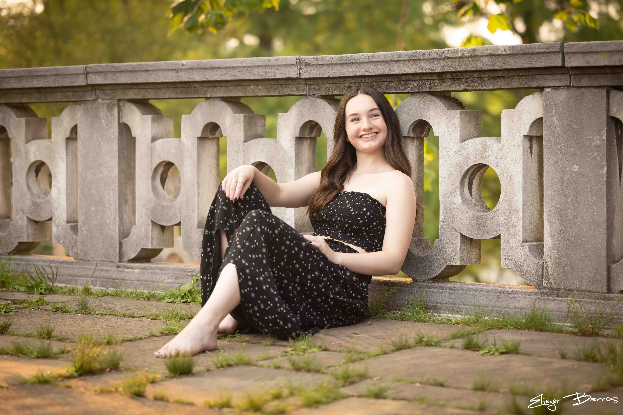 A young woman with long brown hair, smiling, sitting on the ground and leaning against a decorative stone railing in a park with green trees in the background. She is wearing a black dress with white floral patterns.