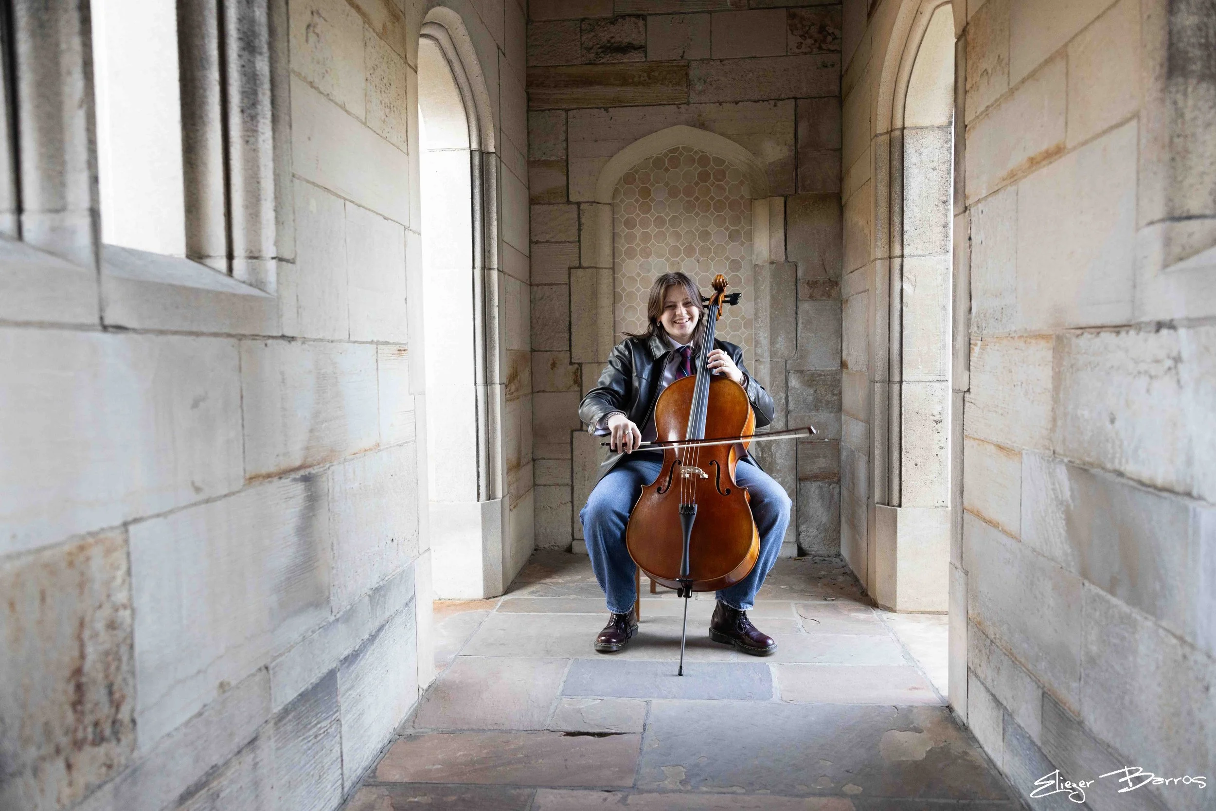 A woman sitting on a chair in a stone corridor playing a cello, wearing a leather jacket and smiling.