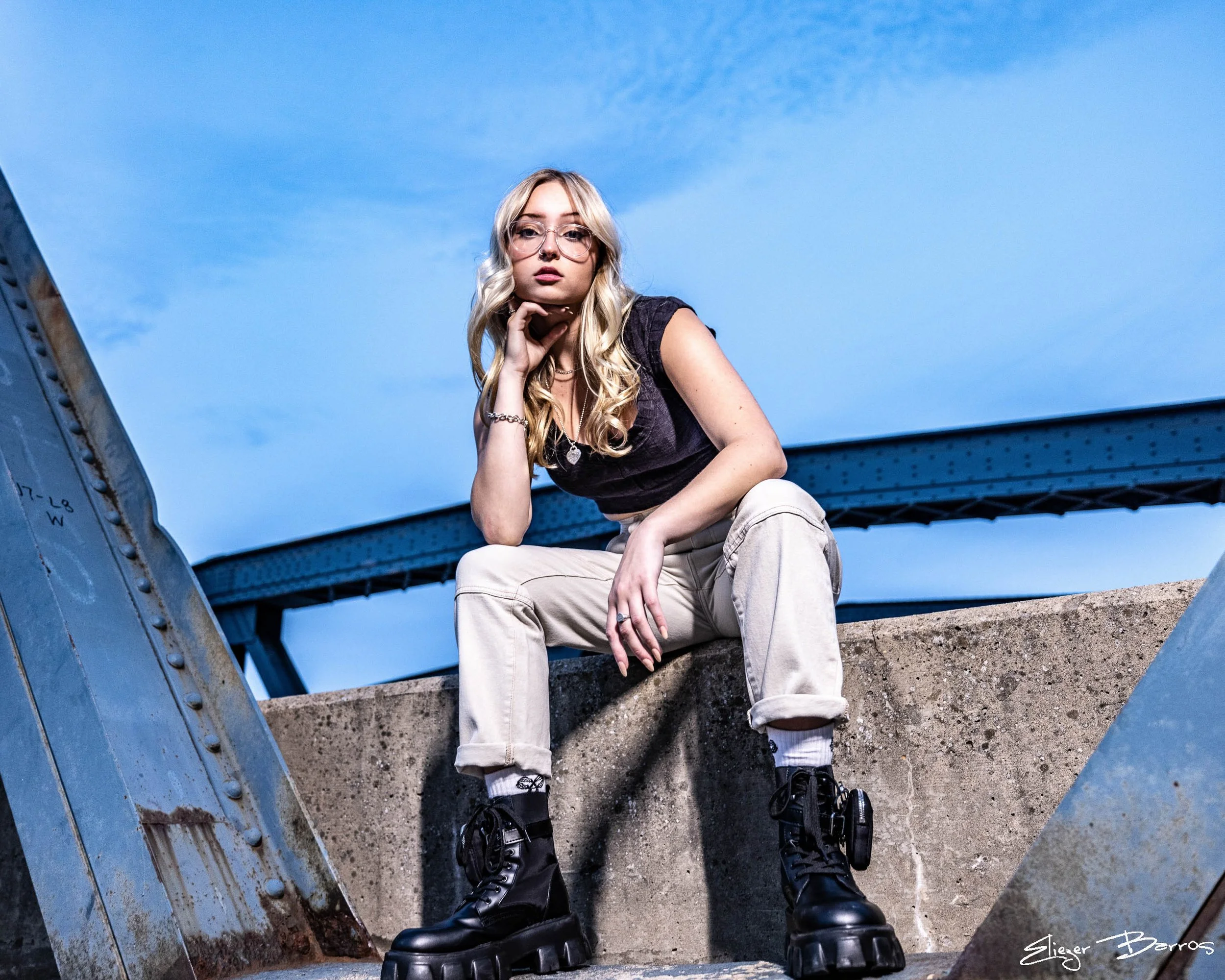 Young woman with blonde hair, glasses, and tattoos, sitting on a concrete ledge beneath a blue bridge during daytime, wearing a black top, beige pants, and black boots.
