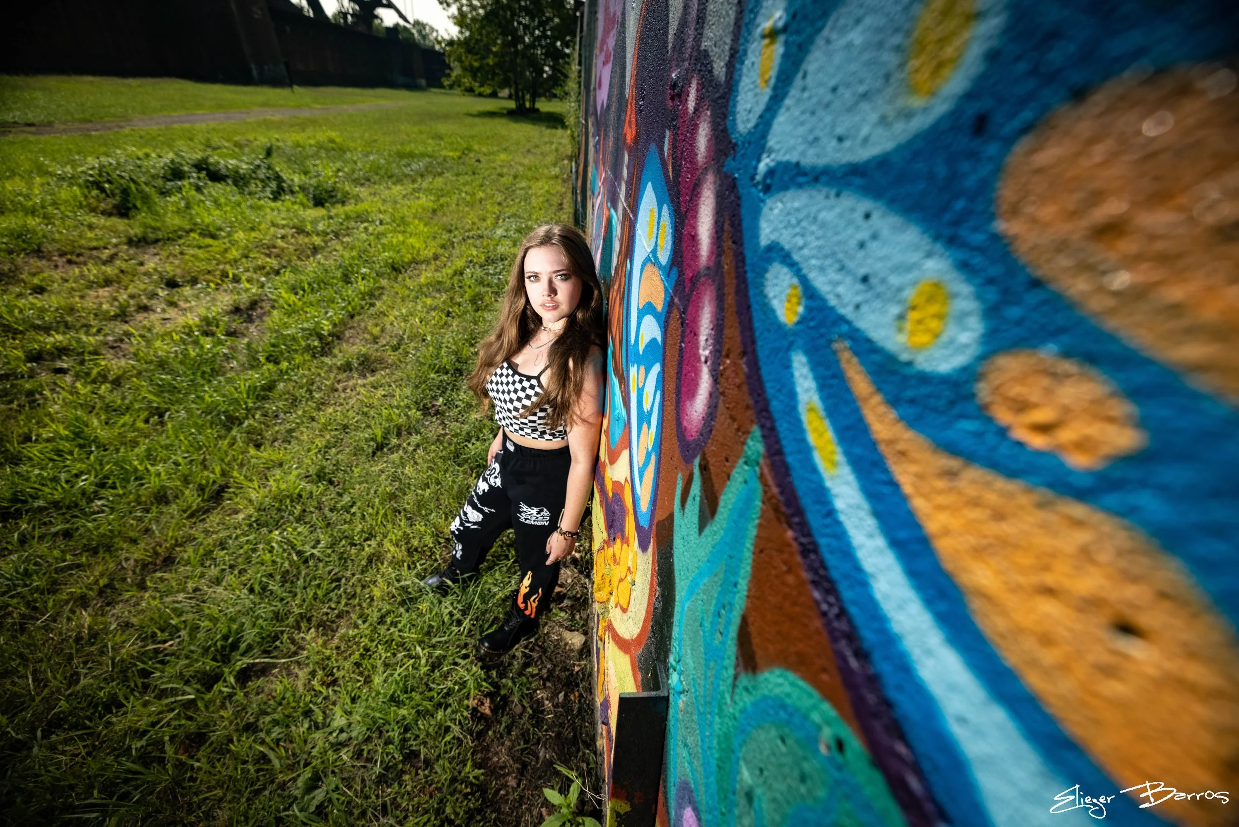 Young woman with long brown hair leaning against a colorful graffiti mural on a brick wall outdoors, with grass and trees in the background.