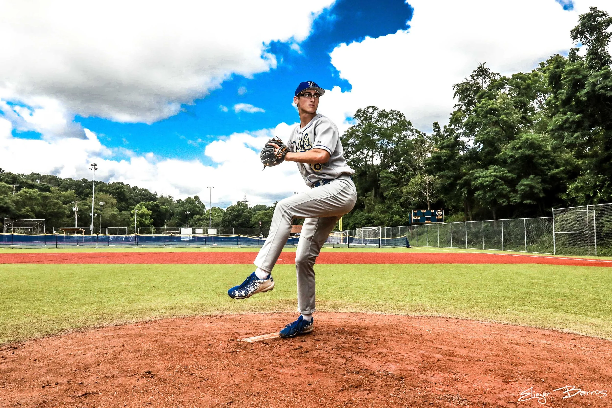 A young baseball pitcher in a gray uniform with a blue cap, standing on a baseball mound, preparing to throw a pitch on a baseball field under a partly cloudy sky, with trees in the background.