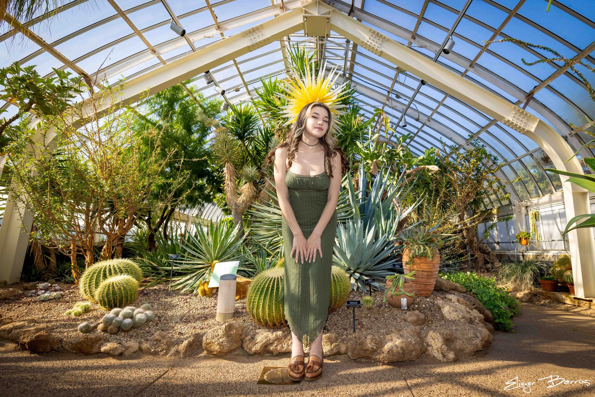 A woman standing in a greenhouse surrounded by various types of cacti and succulents, wearing a green dress with a large yellow spiky plant behind her head, and looking at the camera with a slight smile.