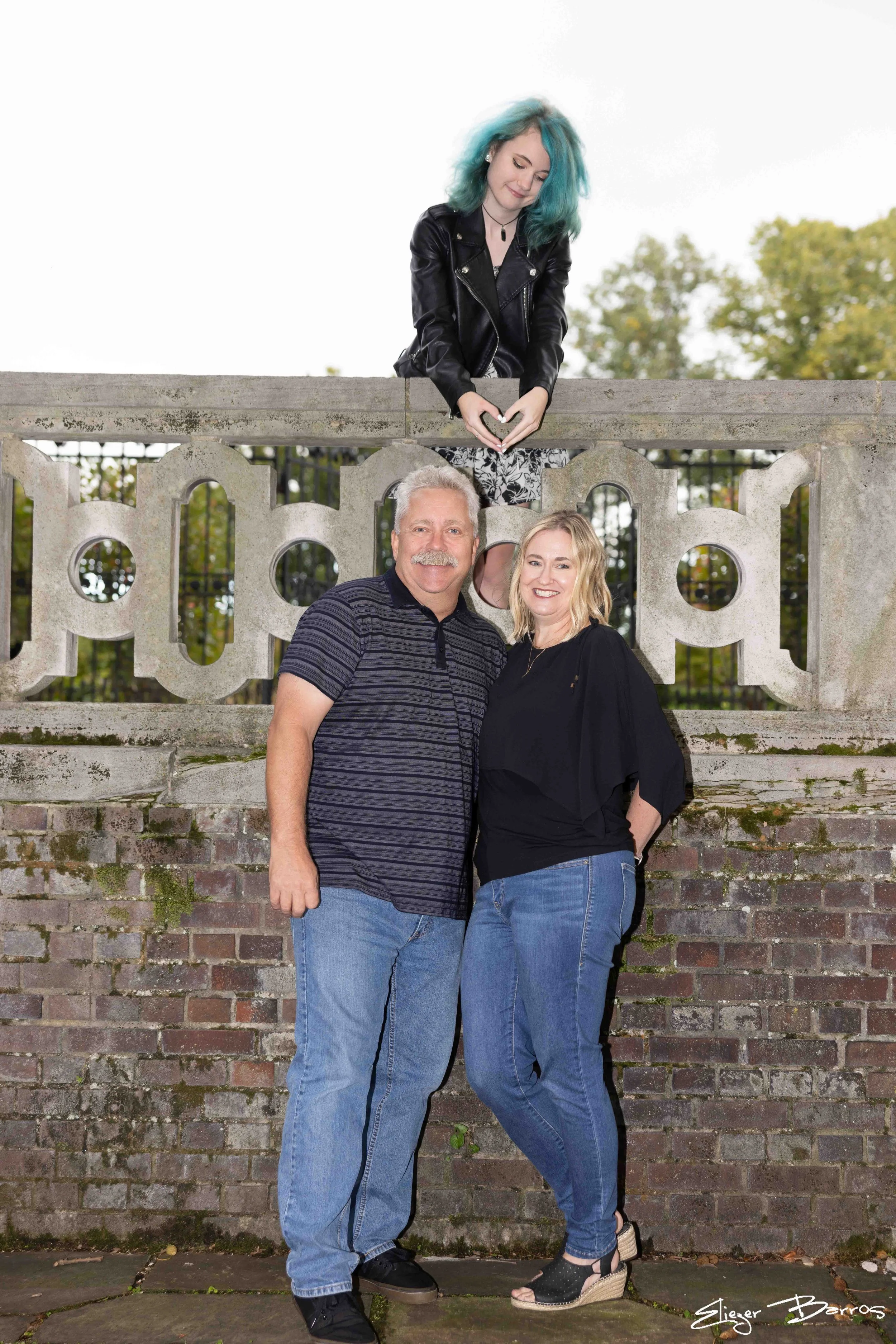 Three people outdoors in front of a concrete wall. A young woman with blue hair, wearing a leather jacket, is on top of the wall making a heart shape with her hands. Two others, a man and a woman, are standing in front of the wall, smiling for the ph