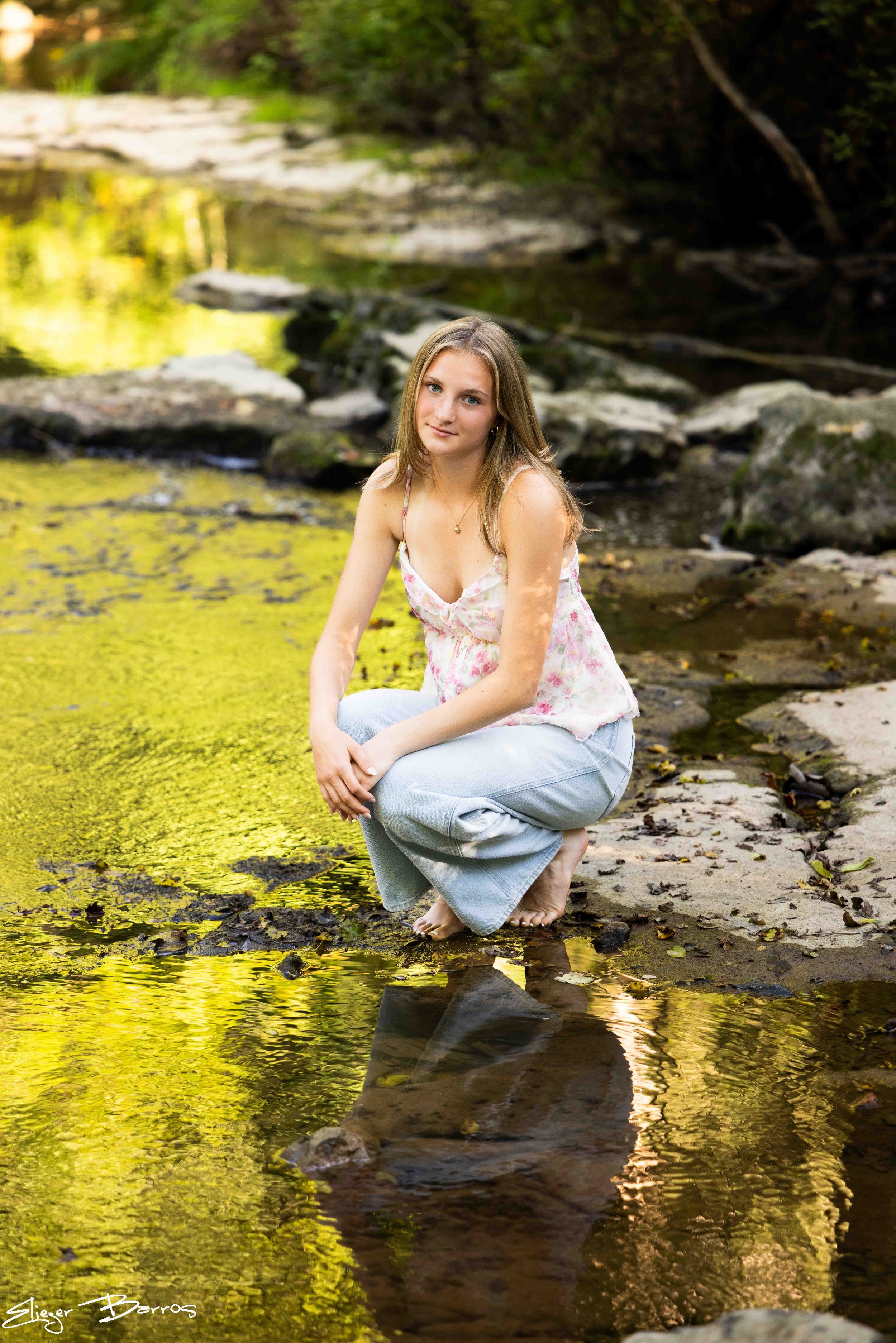 A young woman with long light brown hair, wearing a pastel pink and white sleeveless top and light blue jeans, sitting on a rock by a shallow creek in a forest, with her reflection visible in the water.