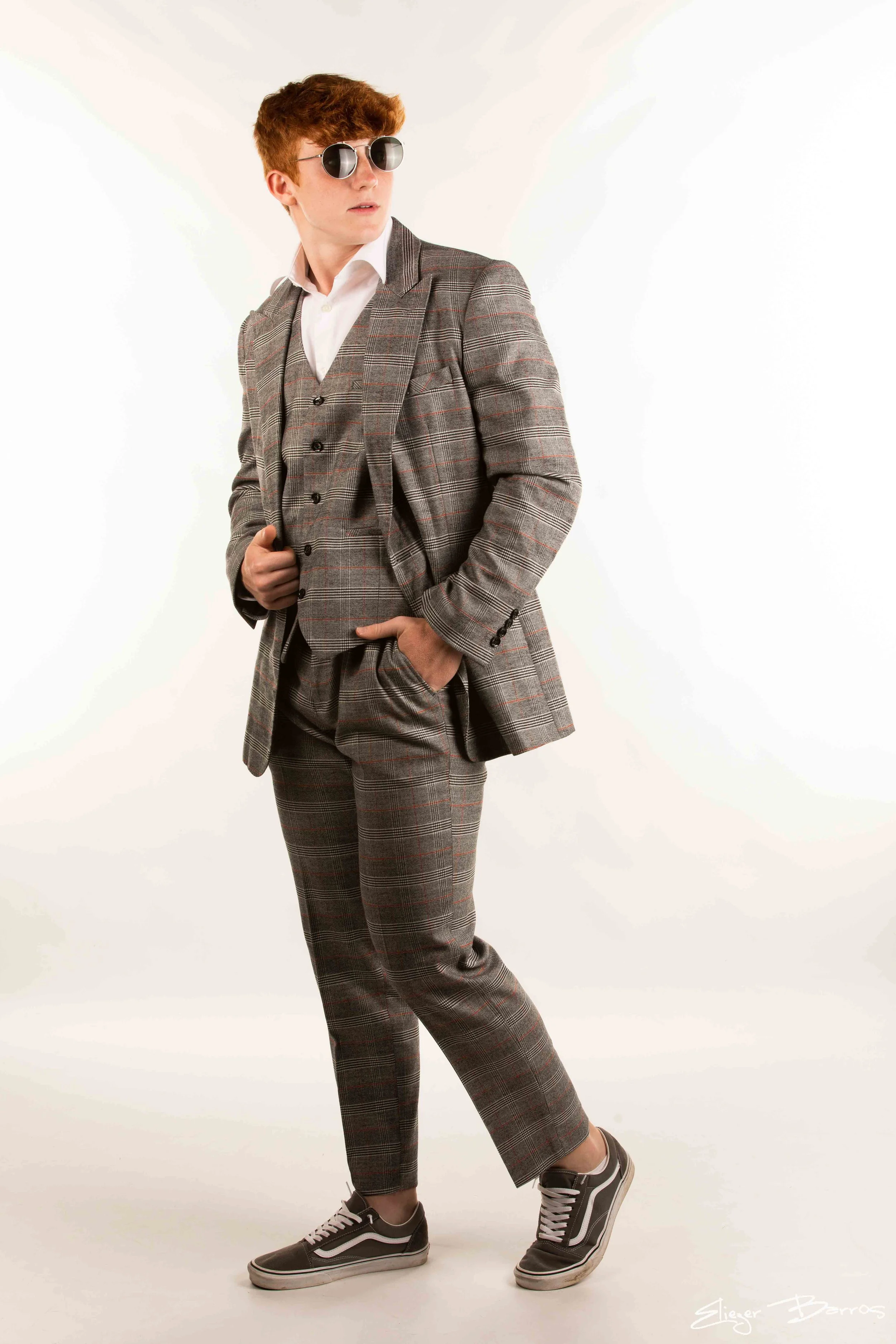 A young man with red hair wearing sunglasses, a gray plaid suit, white shirt, gray sneakers, standing against a plain white background.