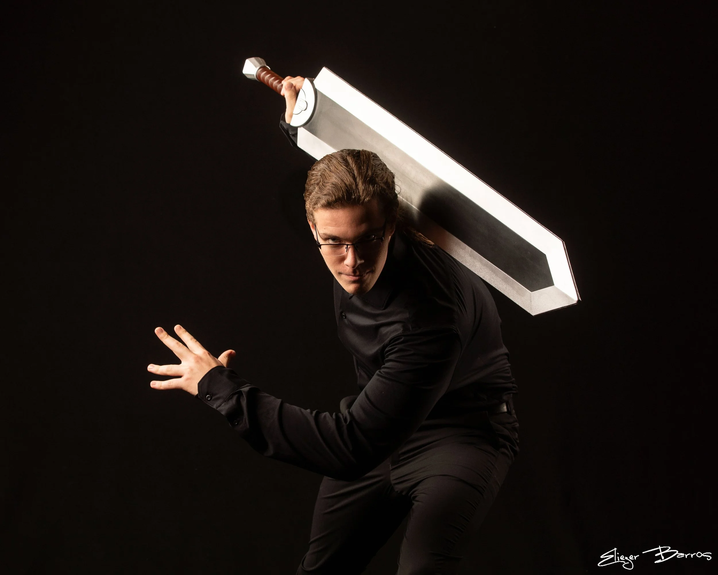 A man wearing glasses in a black shirt holding a large sword over his shoulder against a black background.