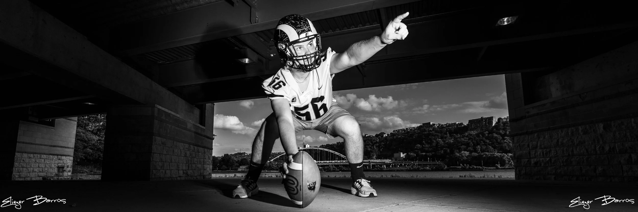 Football player wearing helmet and uniform crouching under bridge, poised to throw a football outdoors with town and bridge in background, black and white photo.