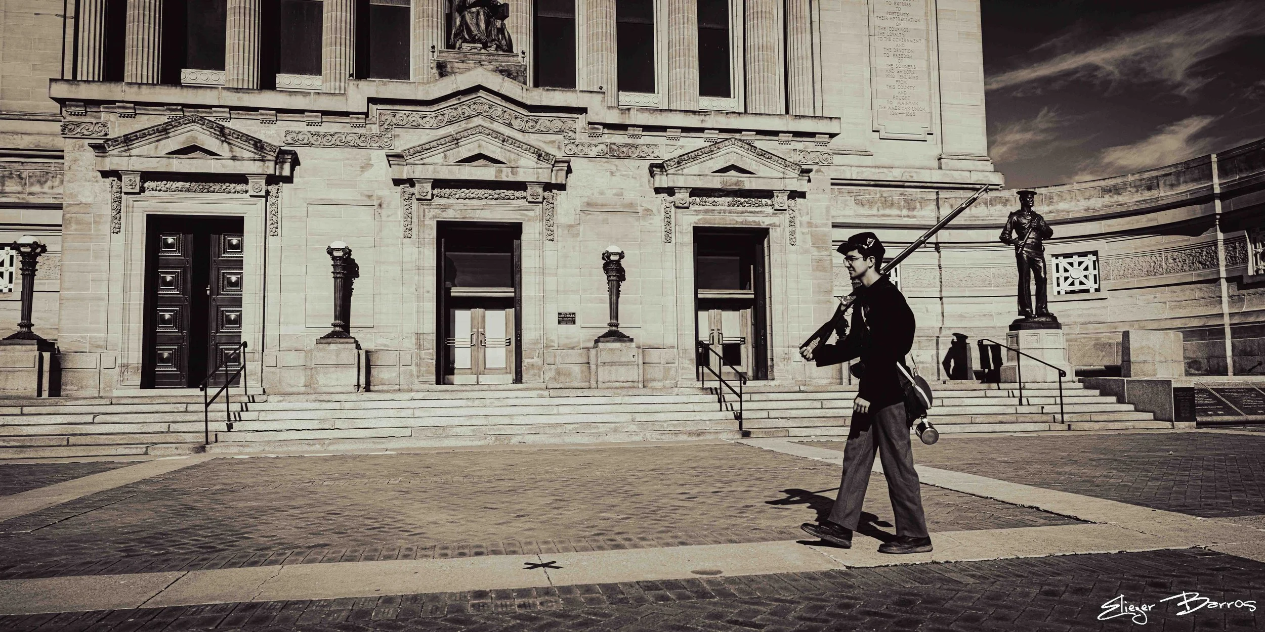 A man carrying a guitar case walking past the steps of a large stone government building with statues and decorative columns.