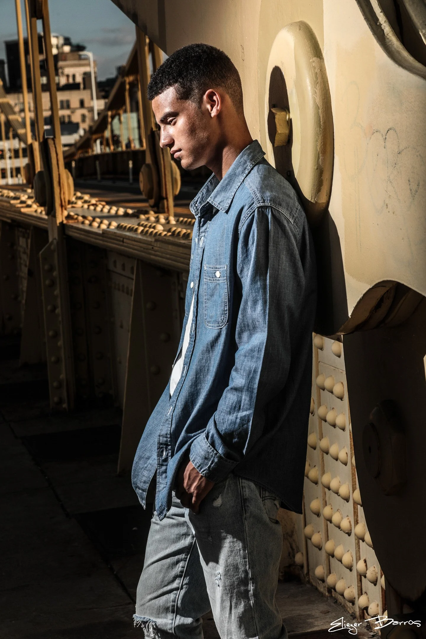 A young man with short curly hair, wearing a denim jacket and ripped jeans, leaning against a wall with a serious expression, in an urban setting with a bridge in the background.
