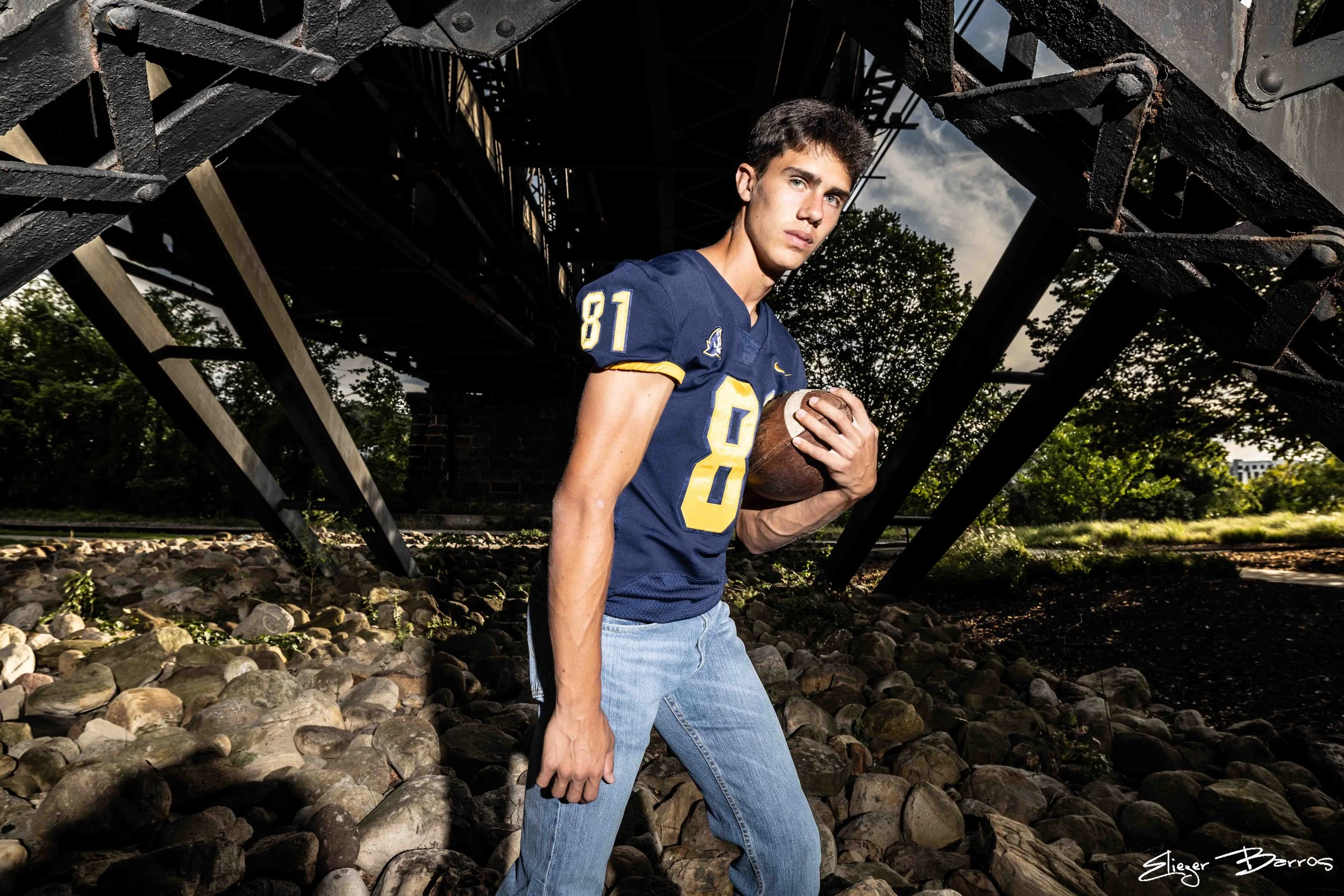 Young man wearing a blue football jersey with number 81, holding a football, standing under a bridge with trees in the background.