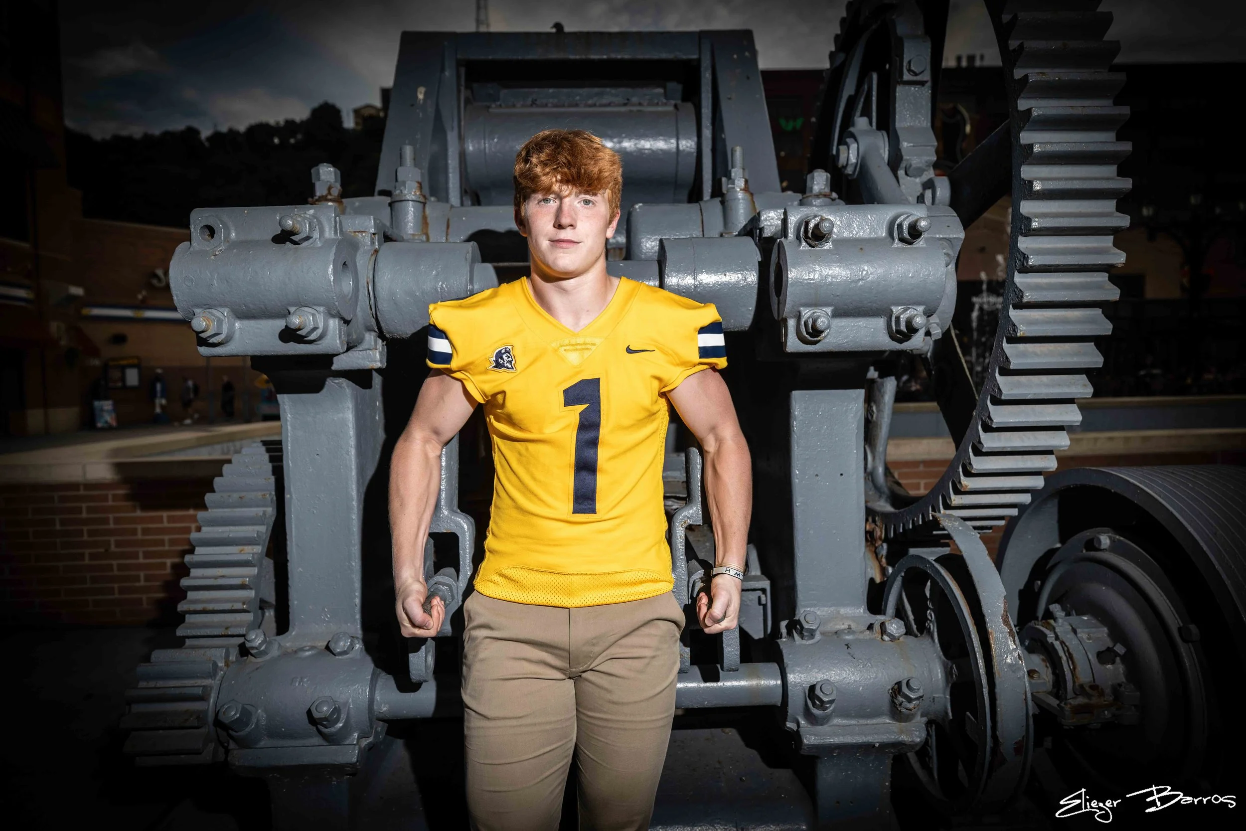 A young man in a yellow football jersey with the number 1 on it, standing in front of a large piece of machinery that resembles a tank tread or industrial equipment.