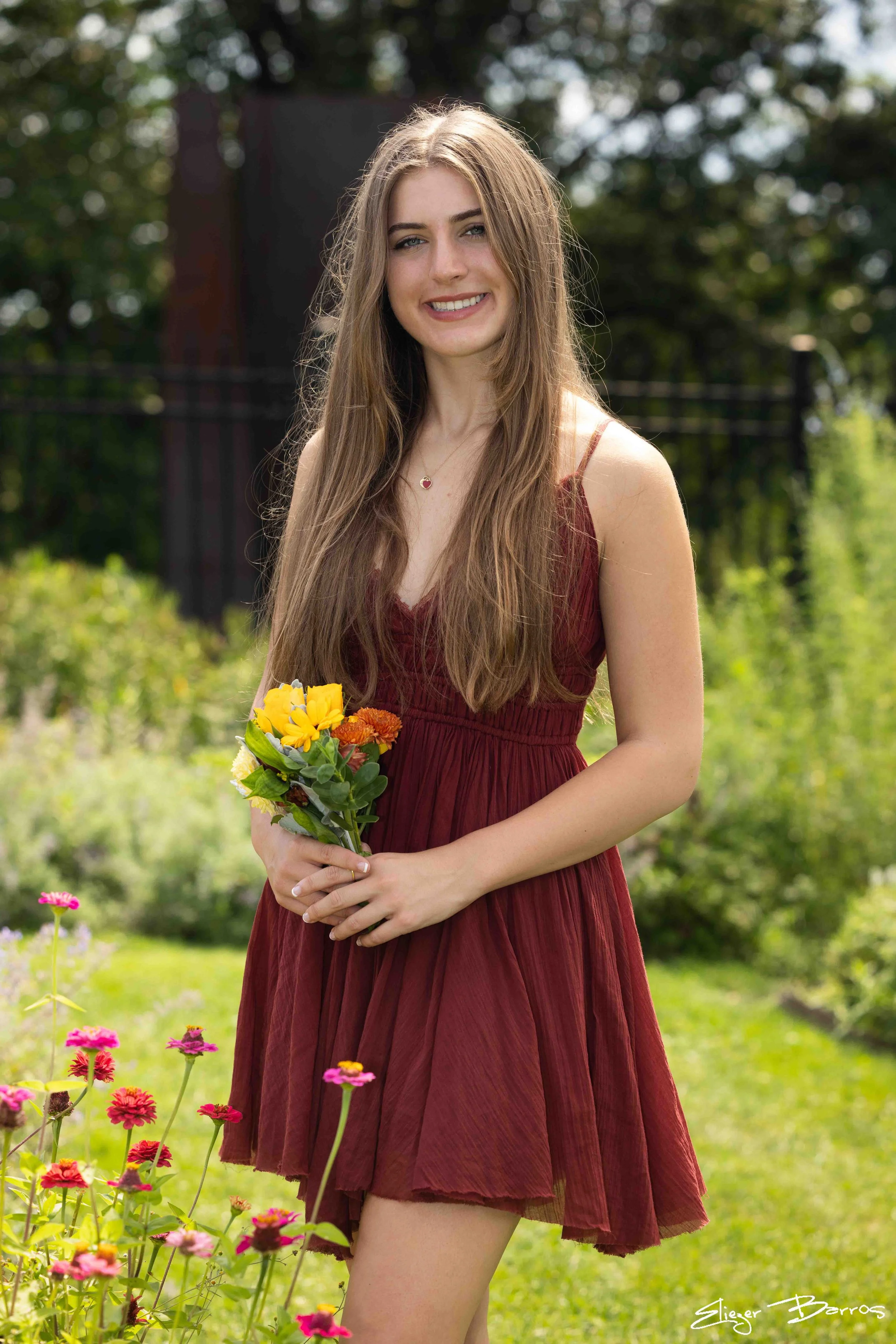 A young woman with long brown hair in a red dress holding a bouquet of yellow and orange flowers standing in a garden with pink and yellow flowers, greenery, and trees in the background.