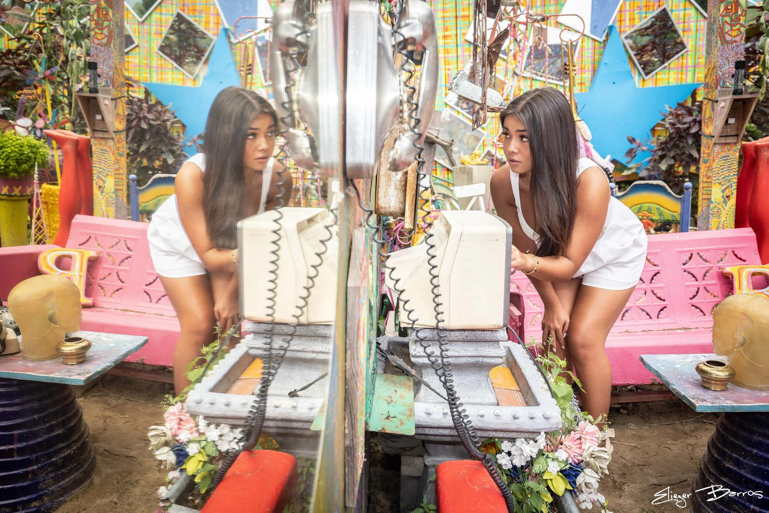 A young woman with long dark hair looking at her reflection in a mirror, photographing herself in a colorful, decorated outdoor setting with Planters, Flowers, and vibrant wall art.