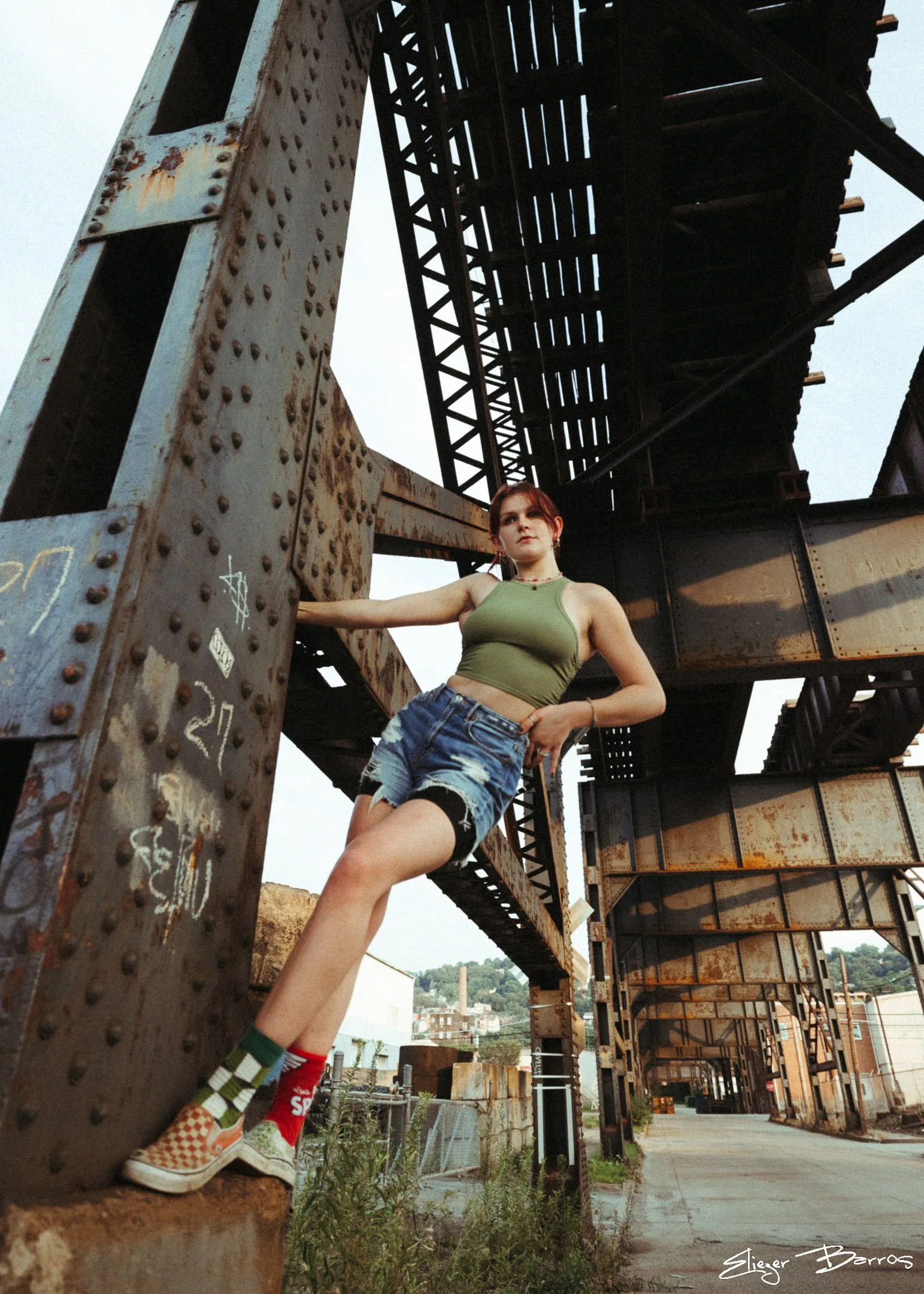 A young woman with red hair in a green tank top and distressed denim shorts leans against a rusty metal structure, part of an old bridge or rail system, with an urban landscape and hillside in the background during the daytime.