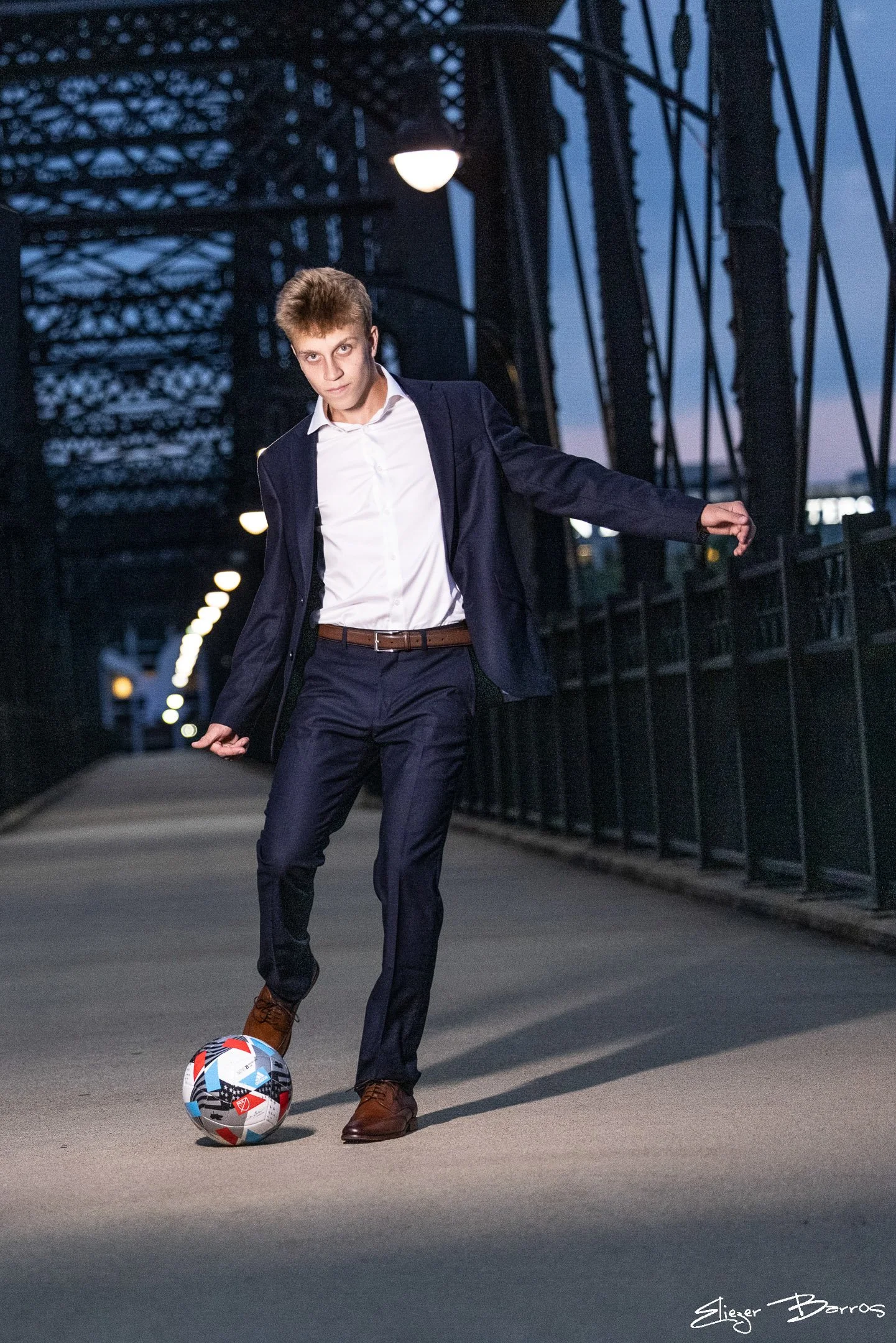 A young man in a business suit playing soccer on a city bridge during dusk. He is dressed in a navy blazer, white shirt, and brown shoes, with a soccer ball at his feet. The city lights are beginning to turn on, illuminating the scene.