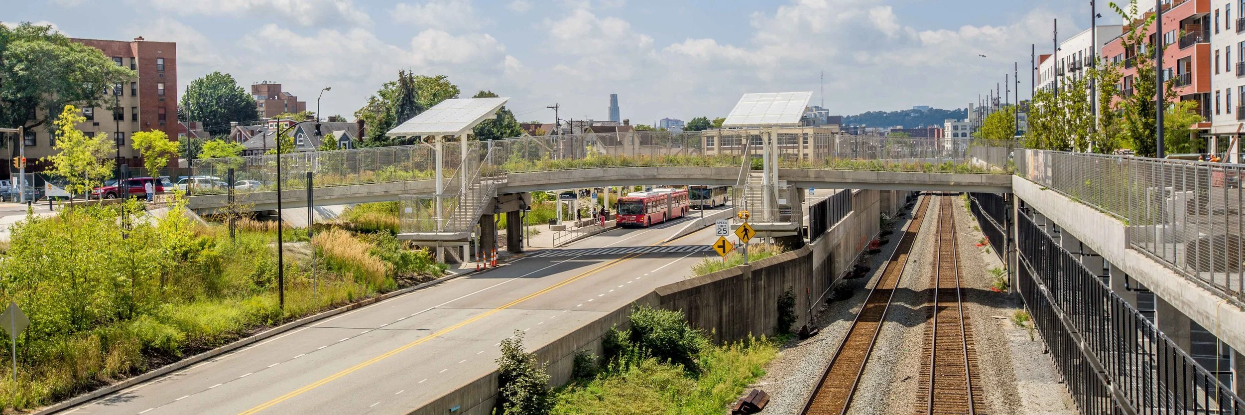 A city scene showing a train station with tracks and a pedestrian bridge, a red bus on the street, and various buildings and trees in the background.