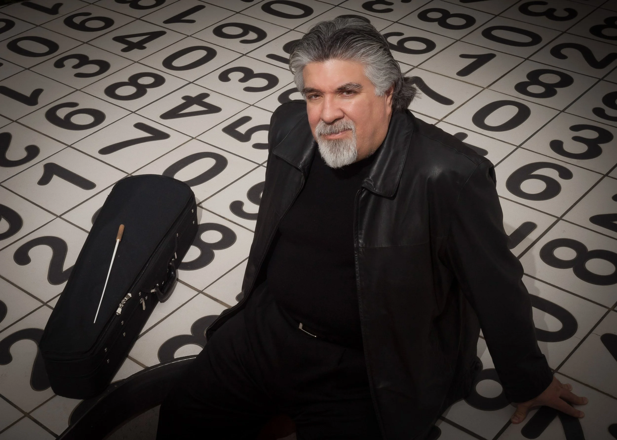 A man with gray hair and a beard wearing a black leather jacket seated on a black stool in front of a floor with large numbered tiles, with a black guitar case beside him.