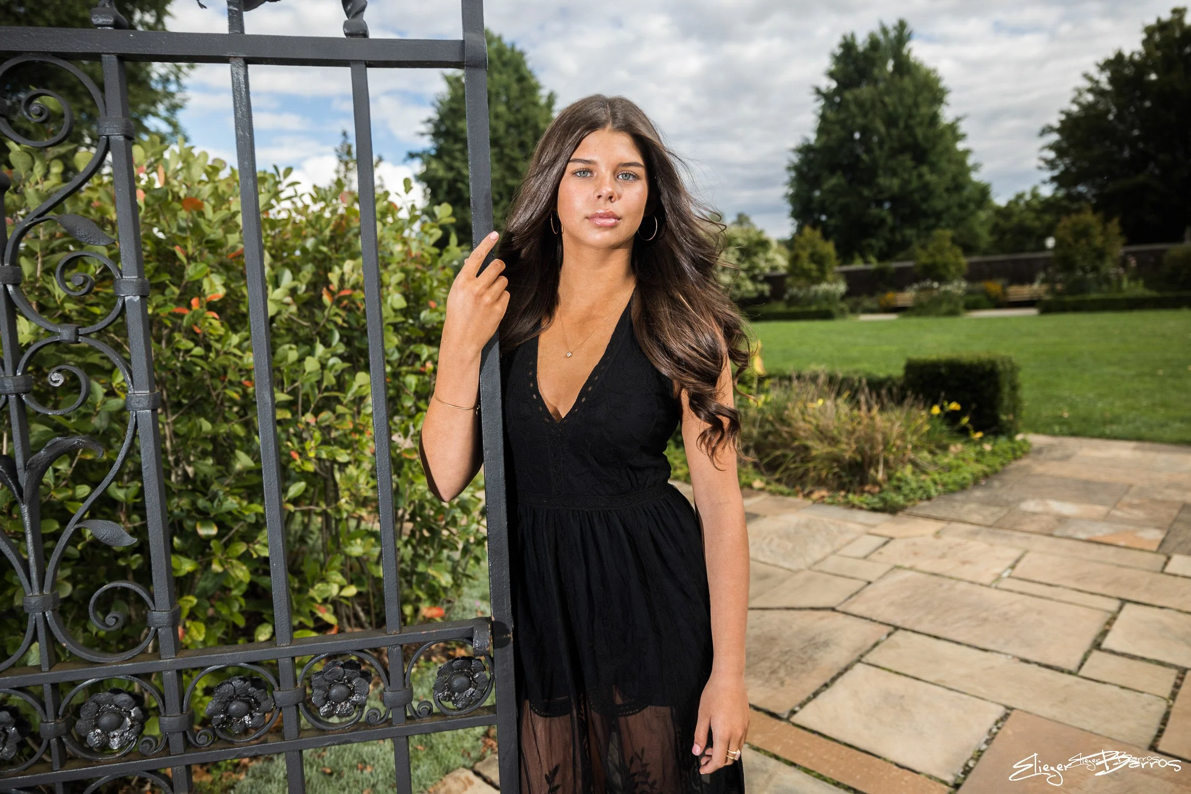Senior Pictures Pittsburgh at Mellon Park. Young woman in a black dress posing by an ornate metal gate in a beautiful garden setting for natural outdoor senior portraits.