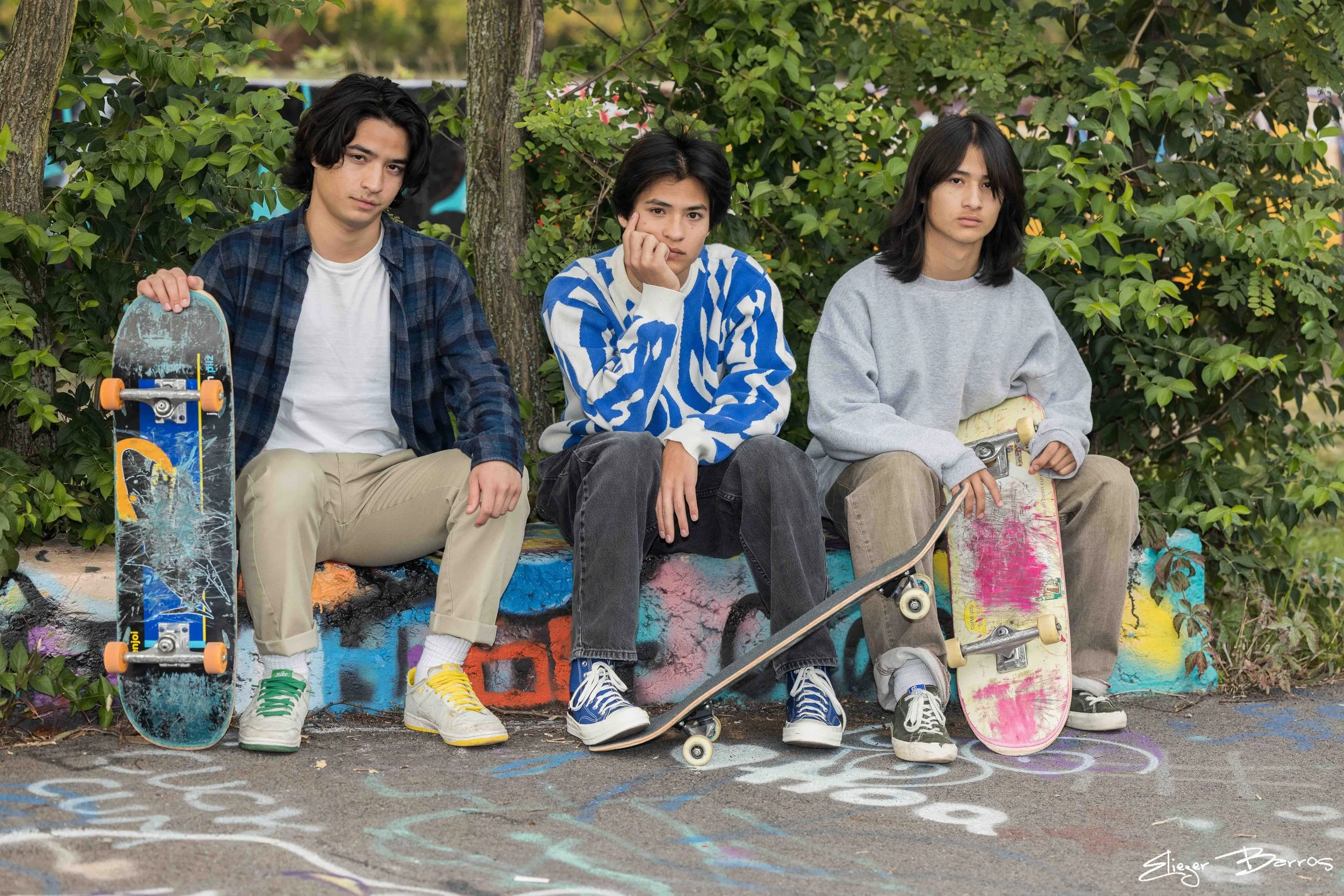 Three young men sitting in front of a graffiti-covered wall and greenery, holding skateboards, dressed casually, with neutral expressions.