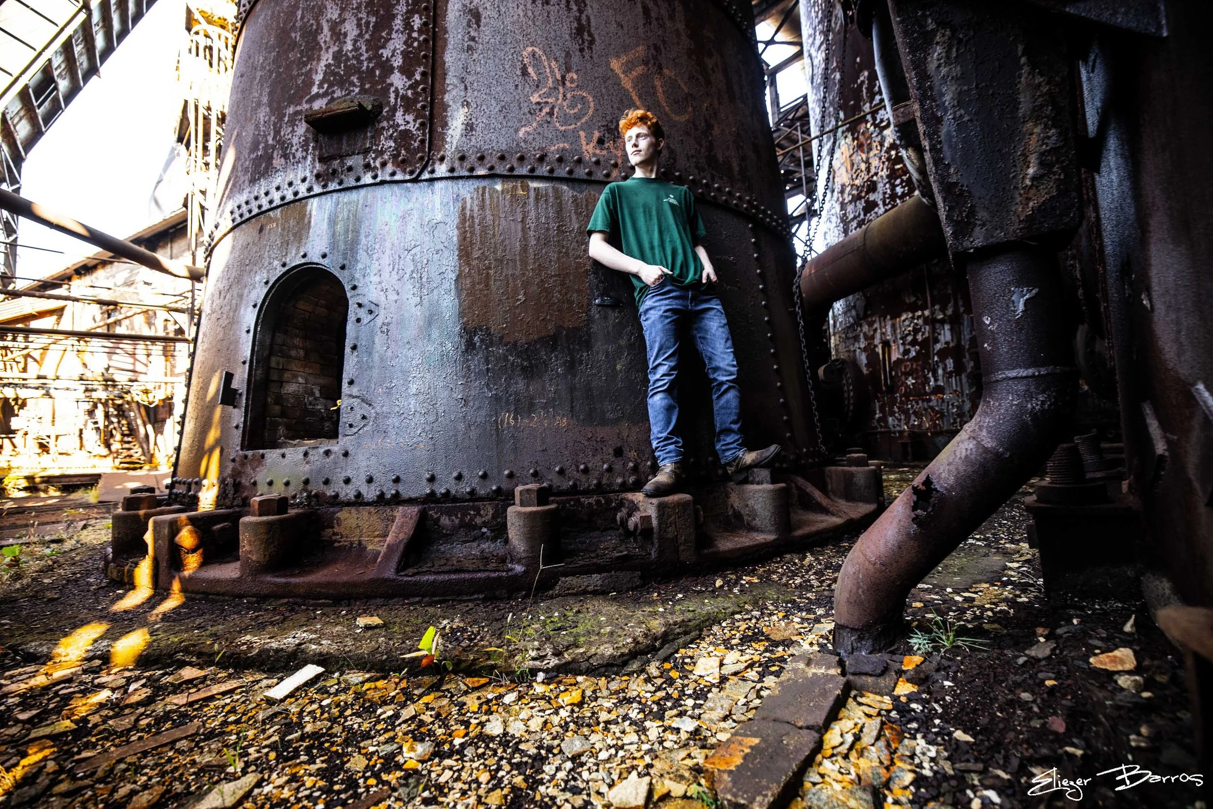 A young man with reddish hair and wearing a dark green T-shirt is standing on a rusty industrial structure, possibly an old factory or refinery, amidst metal pipes and large rusted tanks. The ground is covered with fallen leaves and debris, and the l