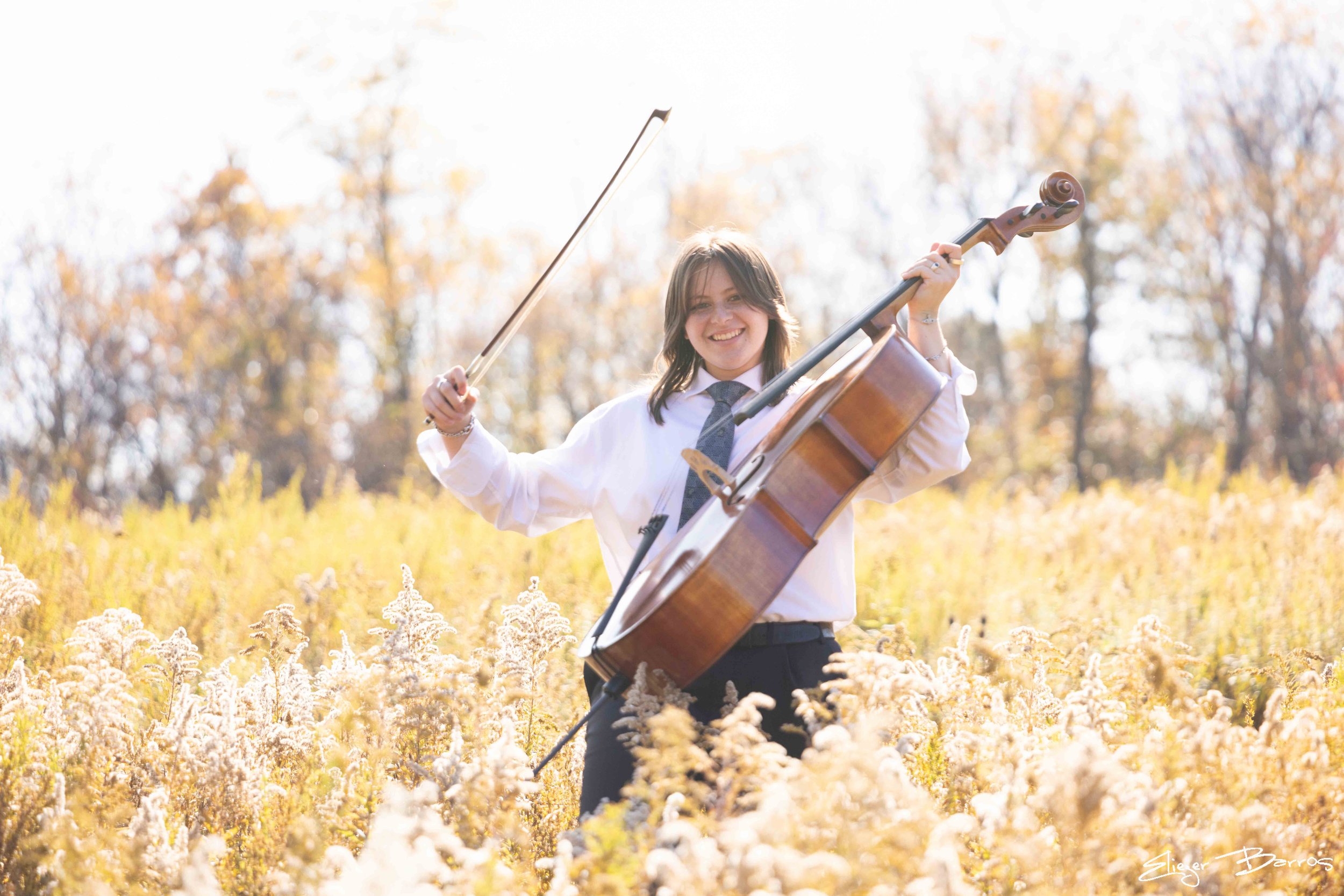 A woman in a white shirt and tie playing a cello outdoors in a field of yellow flowers, with trees in the background during autumn.