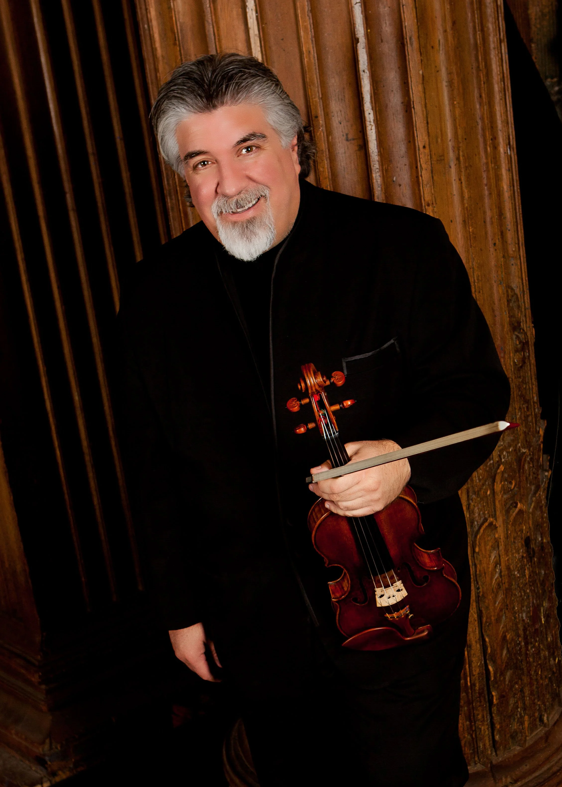 A smiling man with gray hair and a beard, holding a violin and bow, standing in front of a wooden wall.
