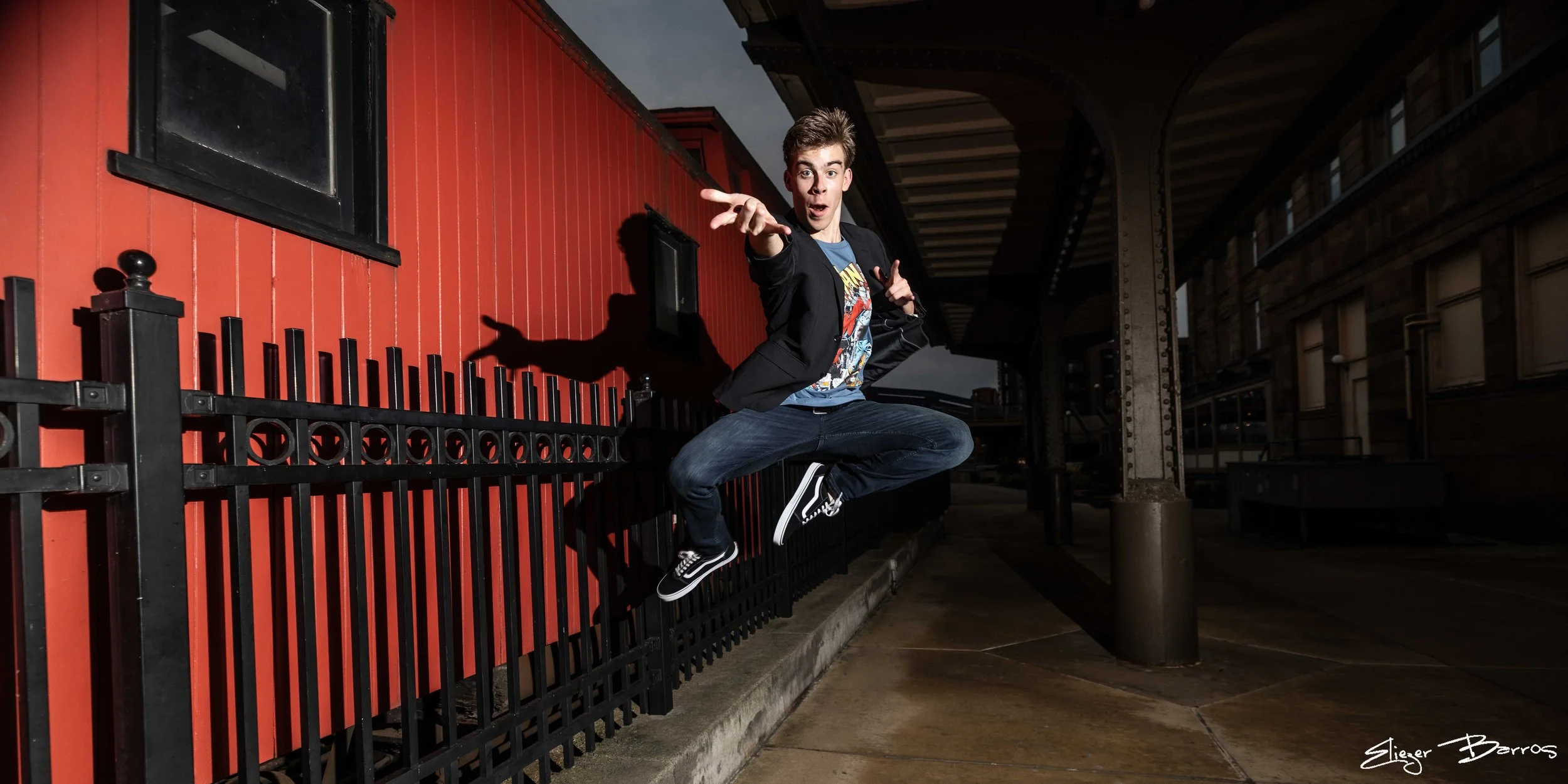A young man wearing a black blazer, blue T-shirt with a graphic design, and jeans is jumping in the air next to a red building with a black fence, during evening or night time in an urban setting.