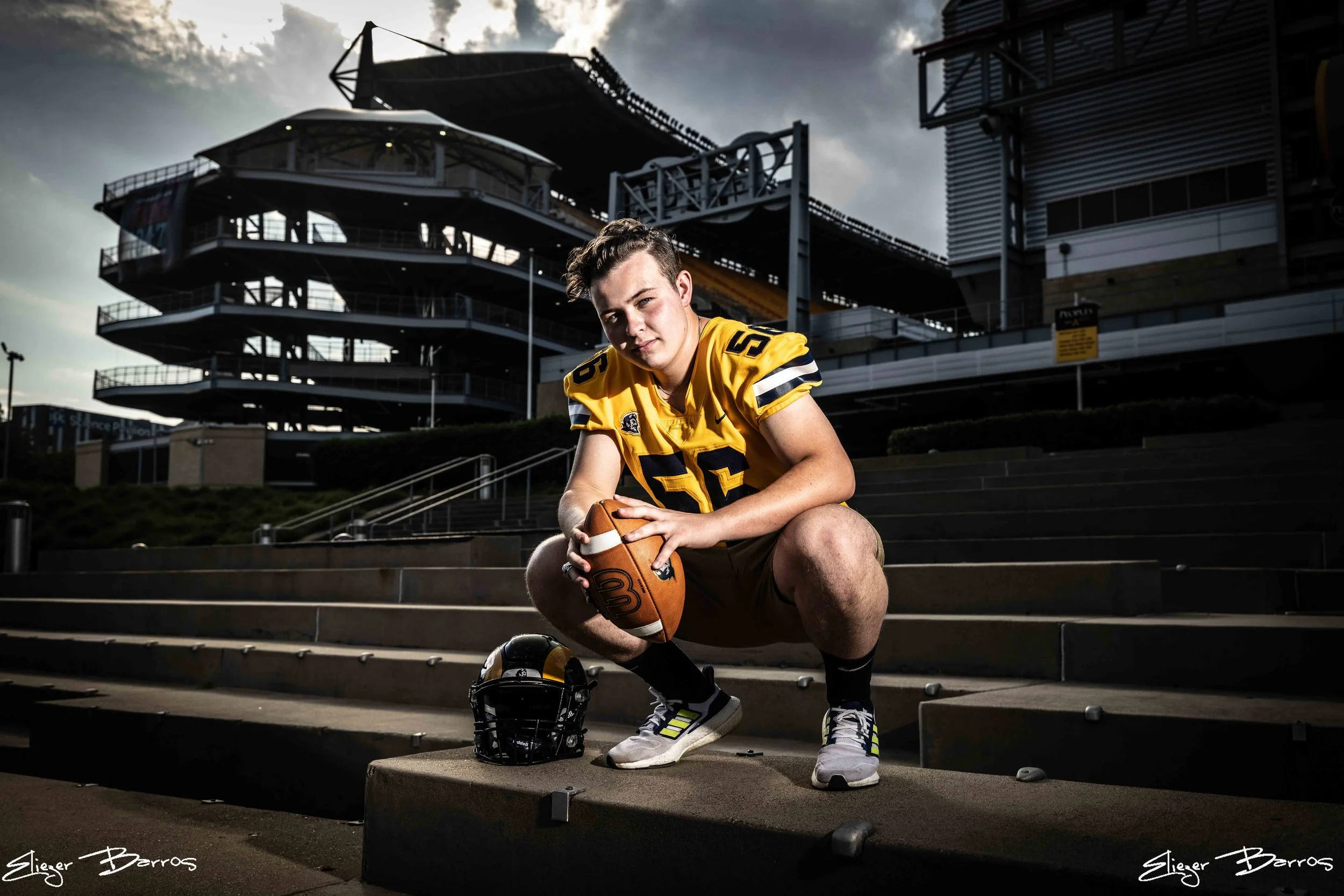 A young male football player in a yellow jersey, holding a football, is kneeling on concrete steps outdoors. His helmet and a pair of shoes are placed on the steps near him. In the background, there is a multi-level parking garage and a building unde
