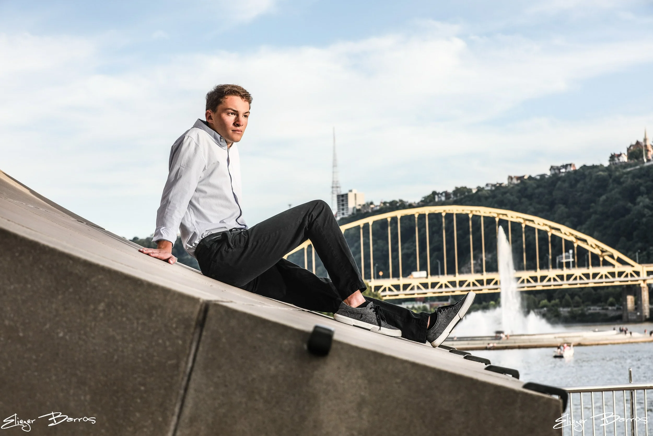 Senior Pictures Pittsburgh at Mister Rogers Memorial. Young man in a white shirt casually sitting on a concrete ledge with fountain and Roberto Clemente Bridge in the background during his scenic senior photoshoot.